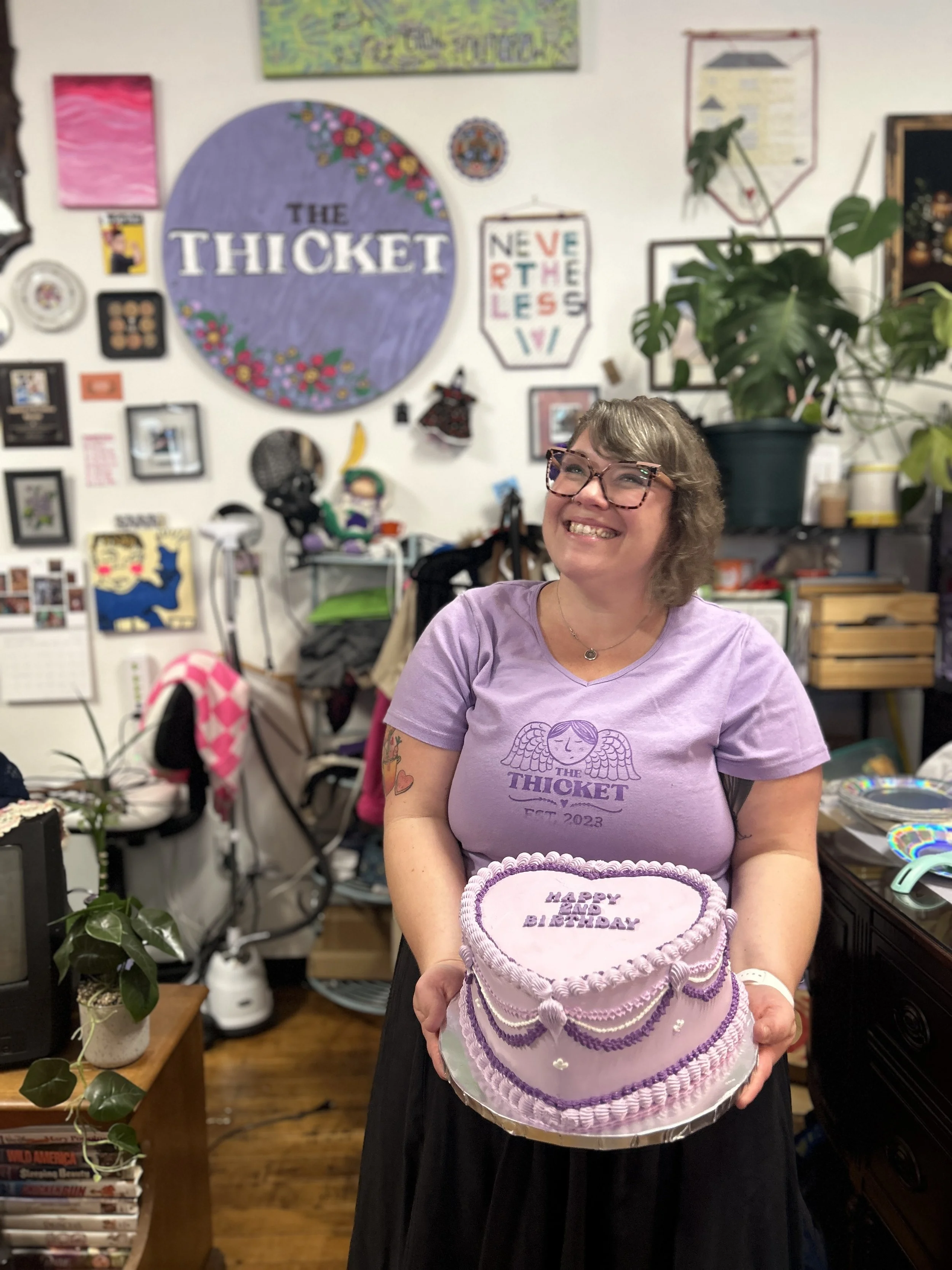 Shop owner, Leigh Soucy, posing in her shop at its 2nd Birthday--she is holding a big heart shaped cake, iced in Thicket purple and is also wearing a Thicket t-shirt.