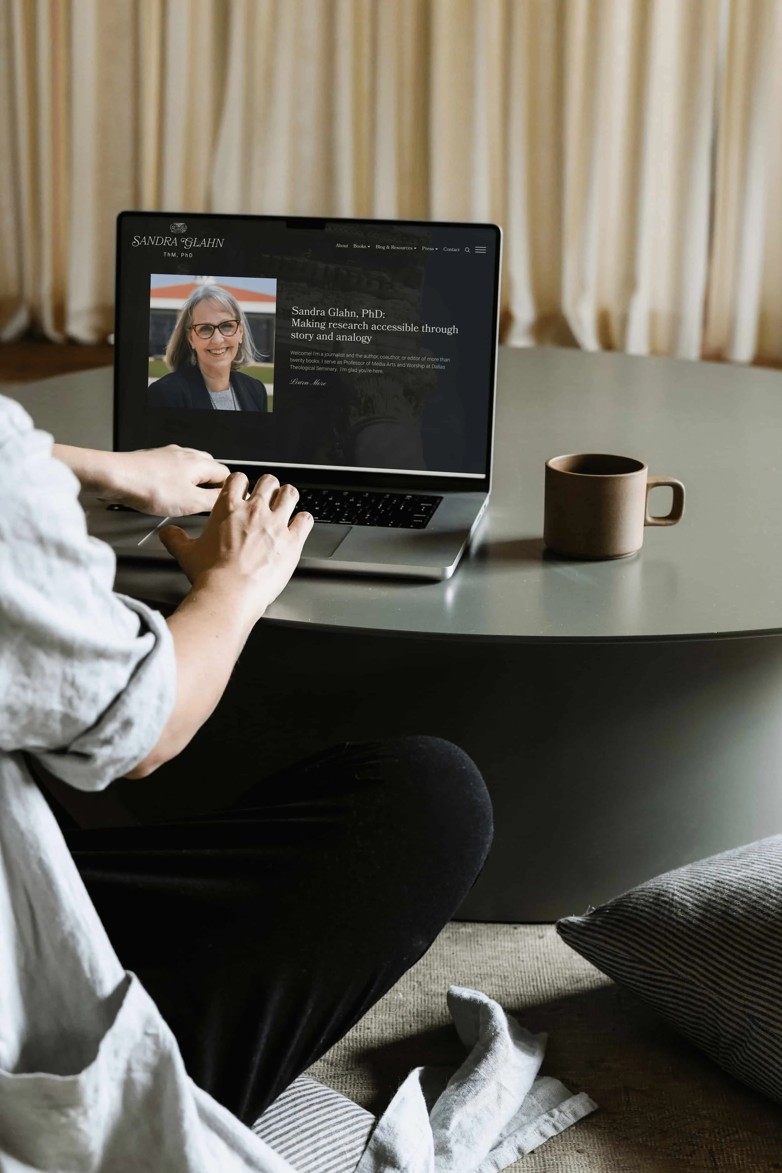 Person typing on a laptop at a table with a mug; laptop screen shows a professional profile of a woman on a website.
