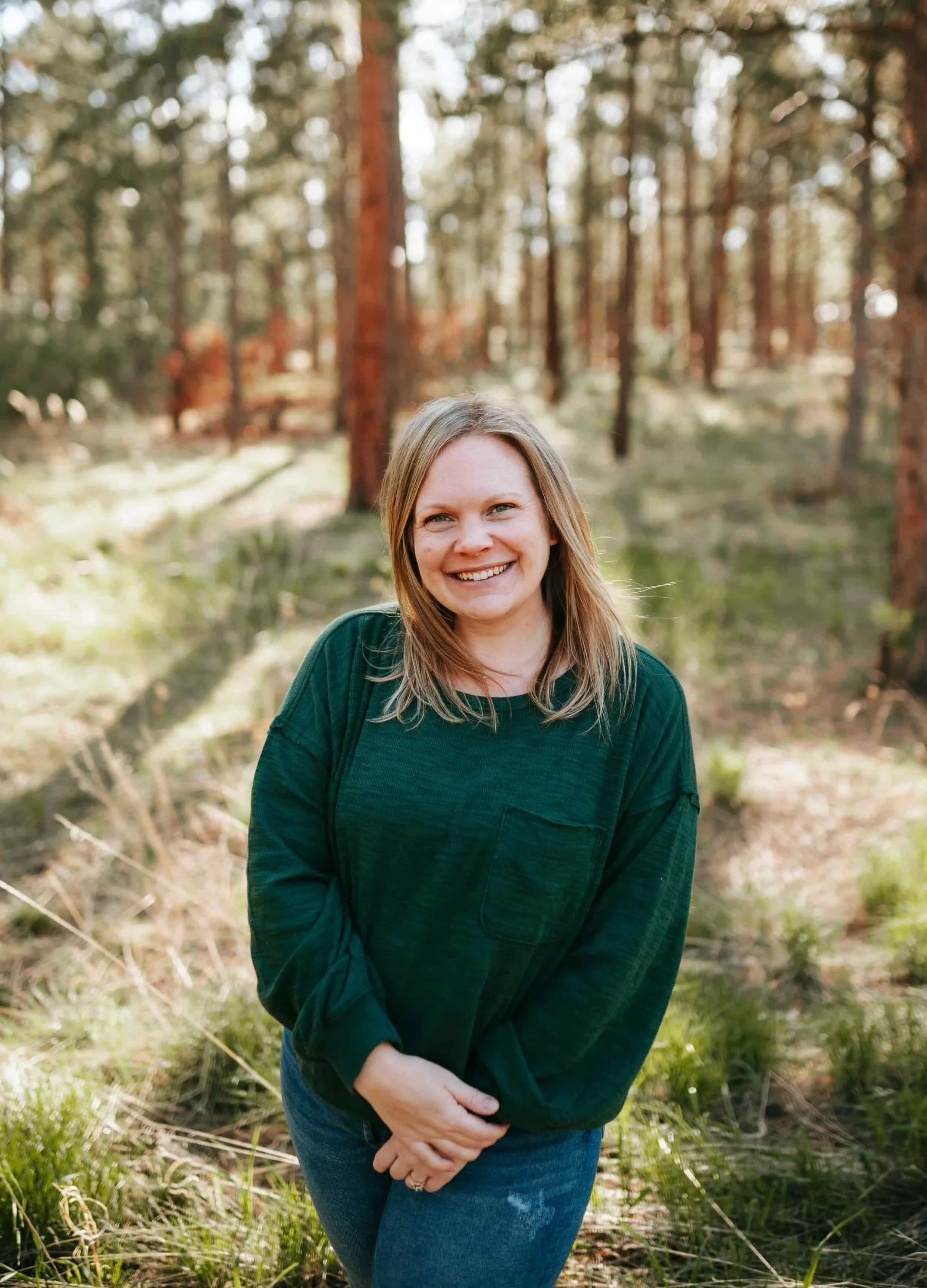 Woman in a green sweater smiling outdoors in a sunlit forest with tall trees and grass.