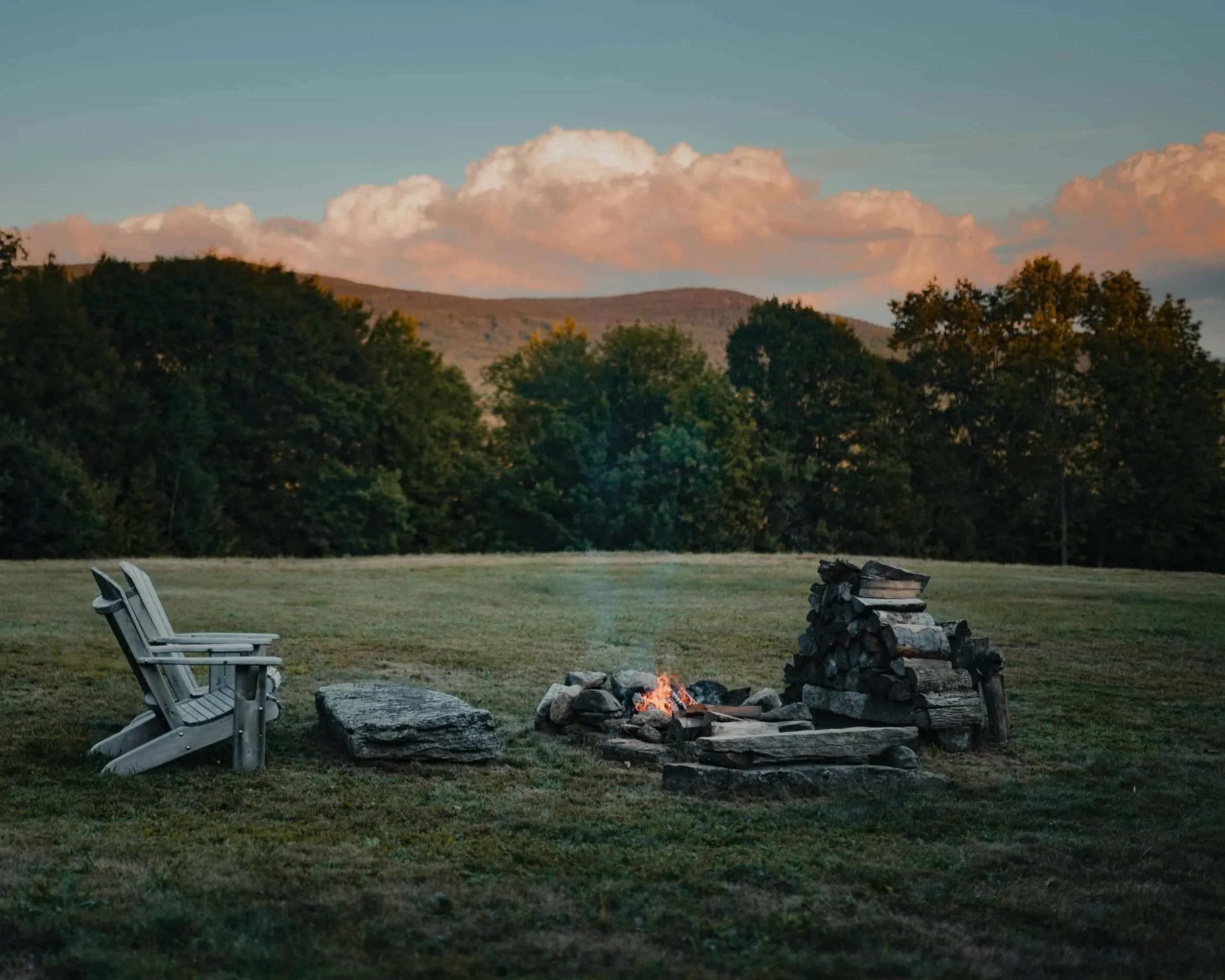 Two empty wooden chairs by a small campfire, surrounded by grass, trees, and distant mountains under a cloudy sky.