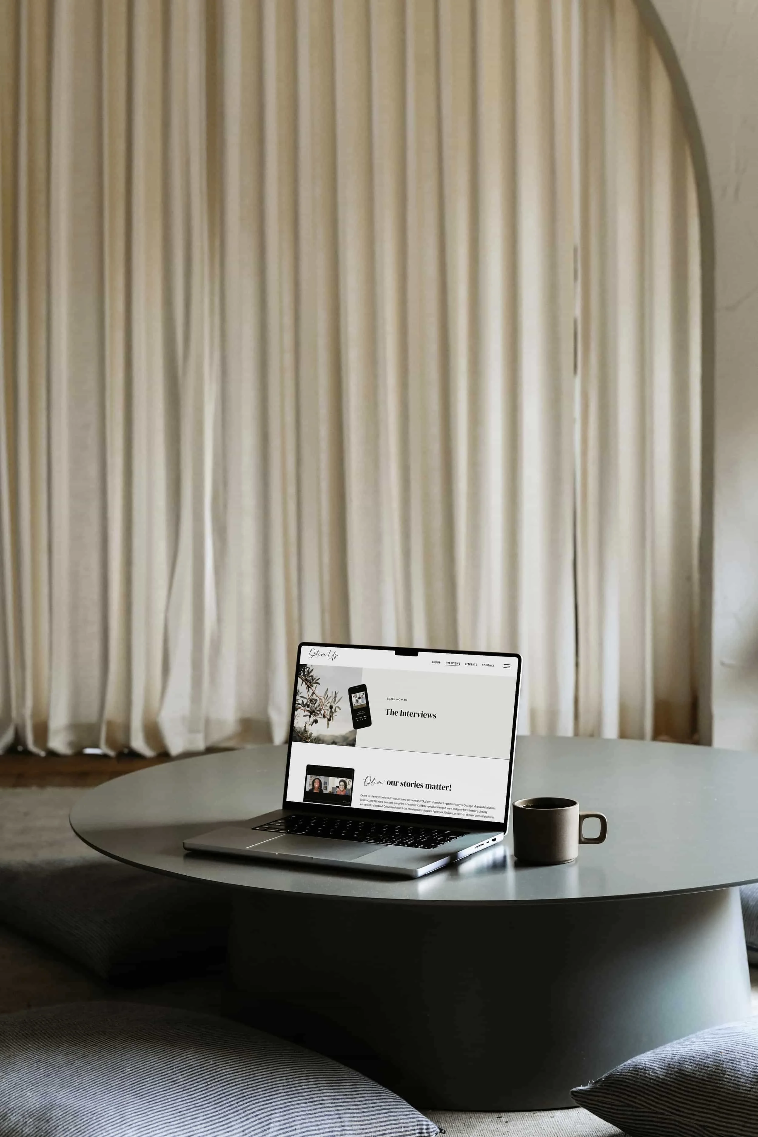 A laptop and a coffee mug sit on a round table in front of large white curtains.