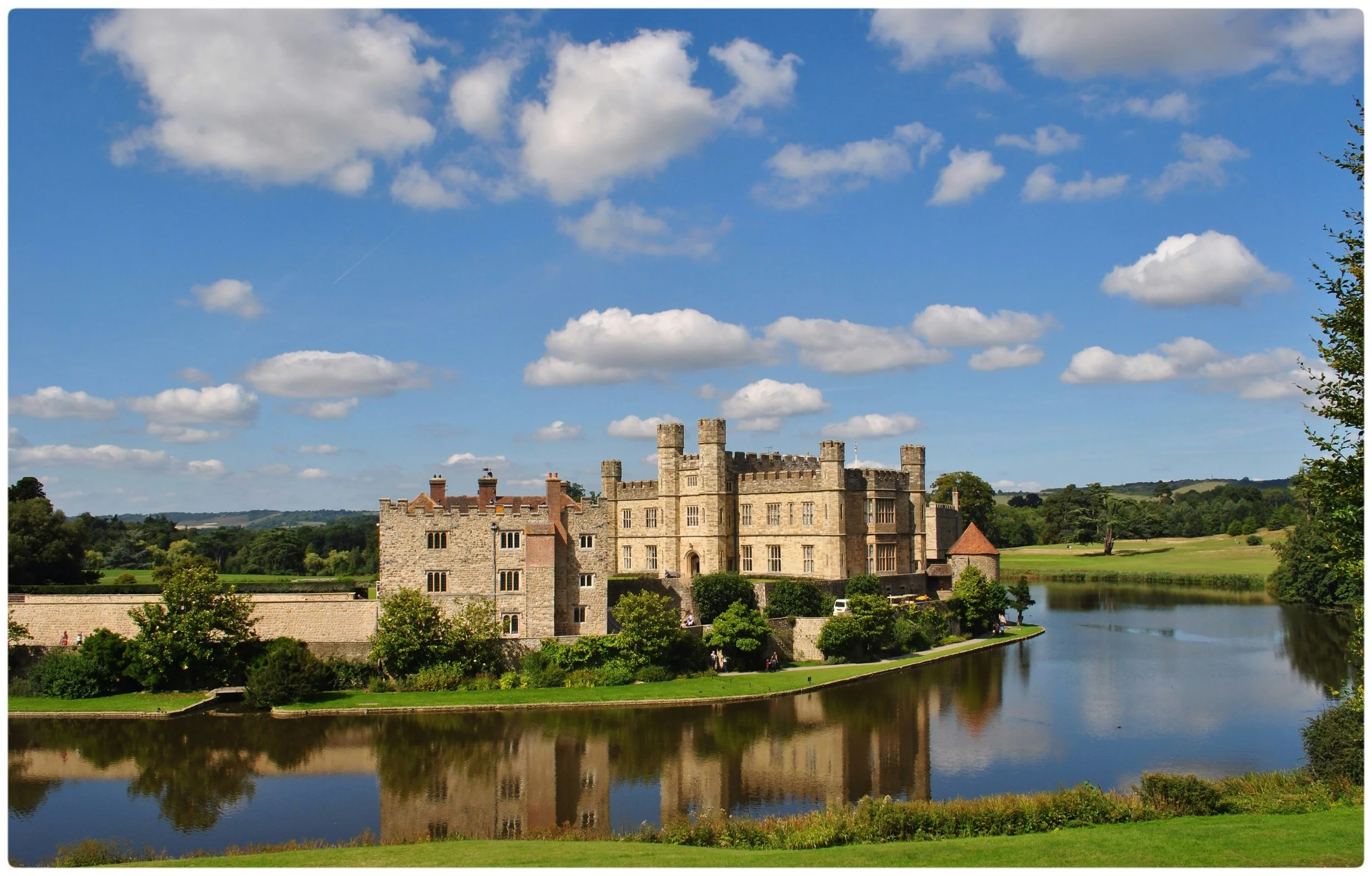 Leeds Castle in Kent reflected in its surrounding lake on a sunny day, with green lawns and rolling English countryside in the background.