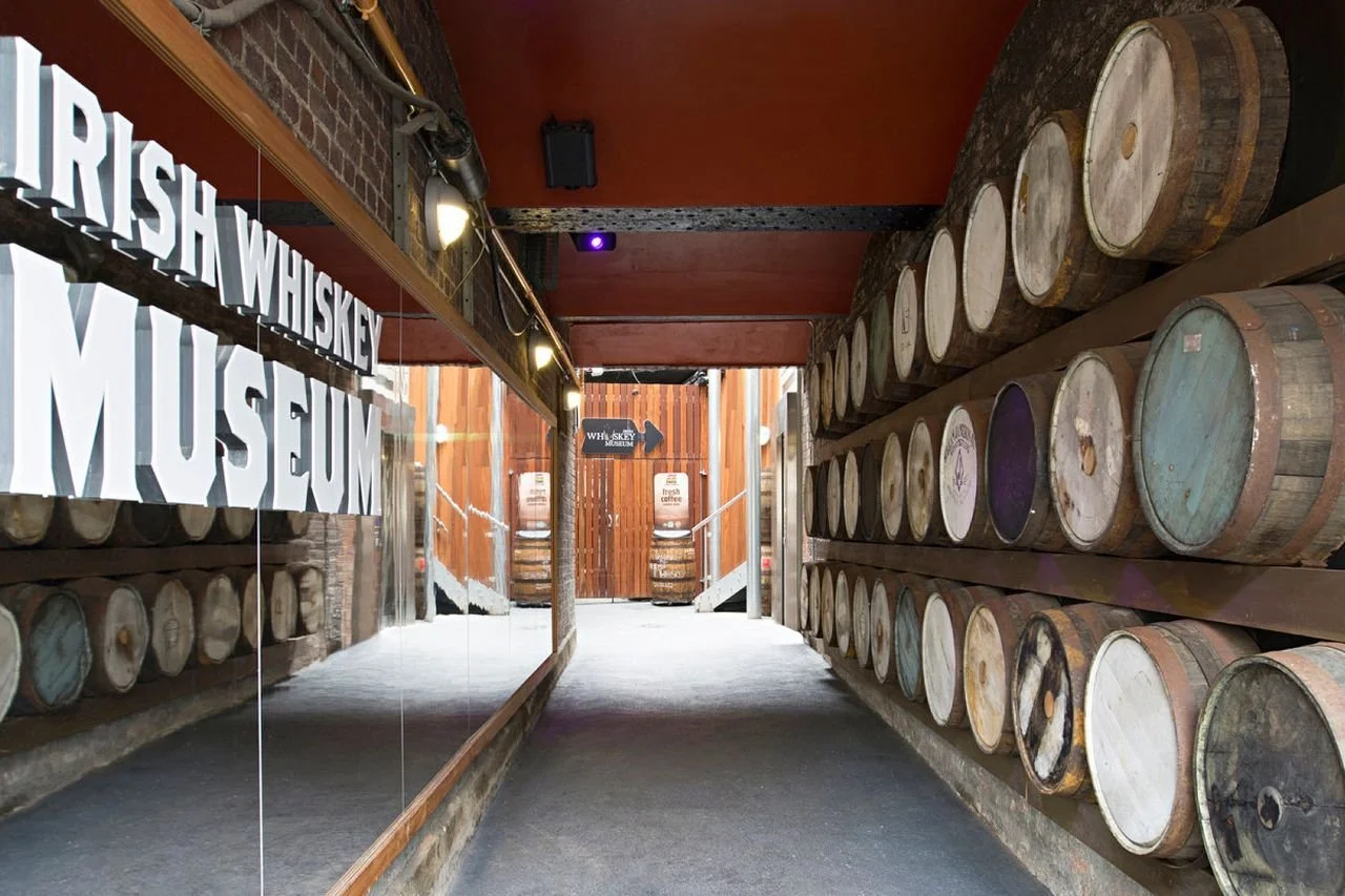 2:56 AM Irish Whiskey Museum entrance corridor lined with stacked wooden whiskey barrels on both sides, illuminated signage reading "Irish Whiskey Museum" on the left wall, leading to wooden doors at the end of the hallway