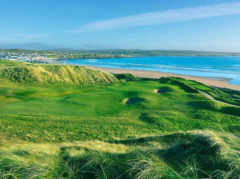 Scenic view of Lahinch Golf Club in County Clare, Ireland, featuring lush green fairways atop coastal dunes overlooking the blue Atlantic Ocean and a sandy beach under a clear sky.