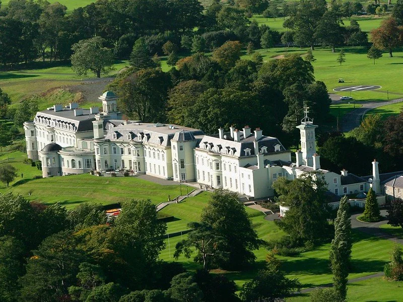 Aerial view of The K Club in County Kildare, Ireland, surrounded by lush green golf courses, trees, and landscaped gardens under clear skies.