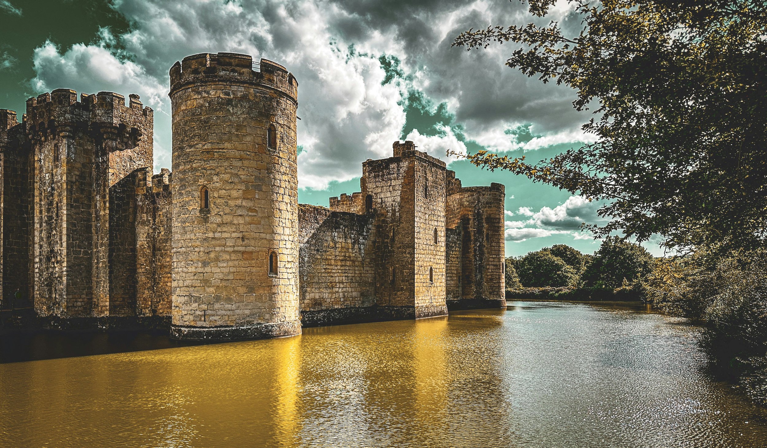 Bodiam Castle in East Sussex surrounded by a wide reflective moat, its stone towers rising under dramatic clouds and framed by trees.