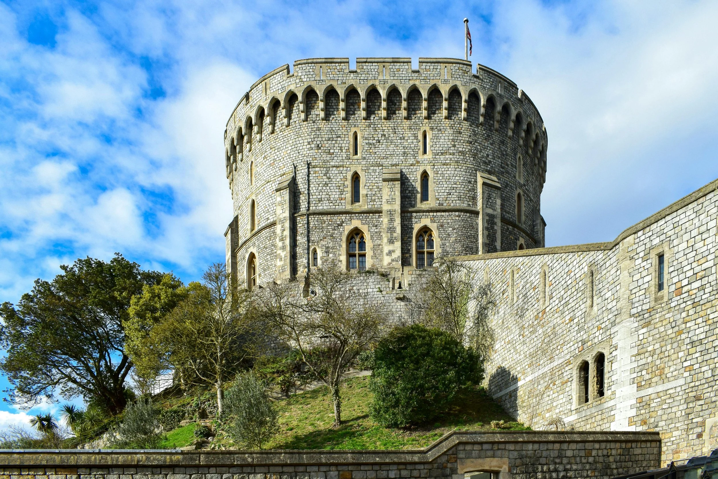 The Round Tower of Windsor Castle rising above stone walls and trees under a bright blue sky in England.