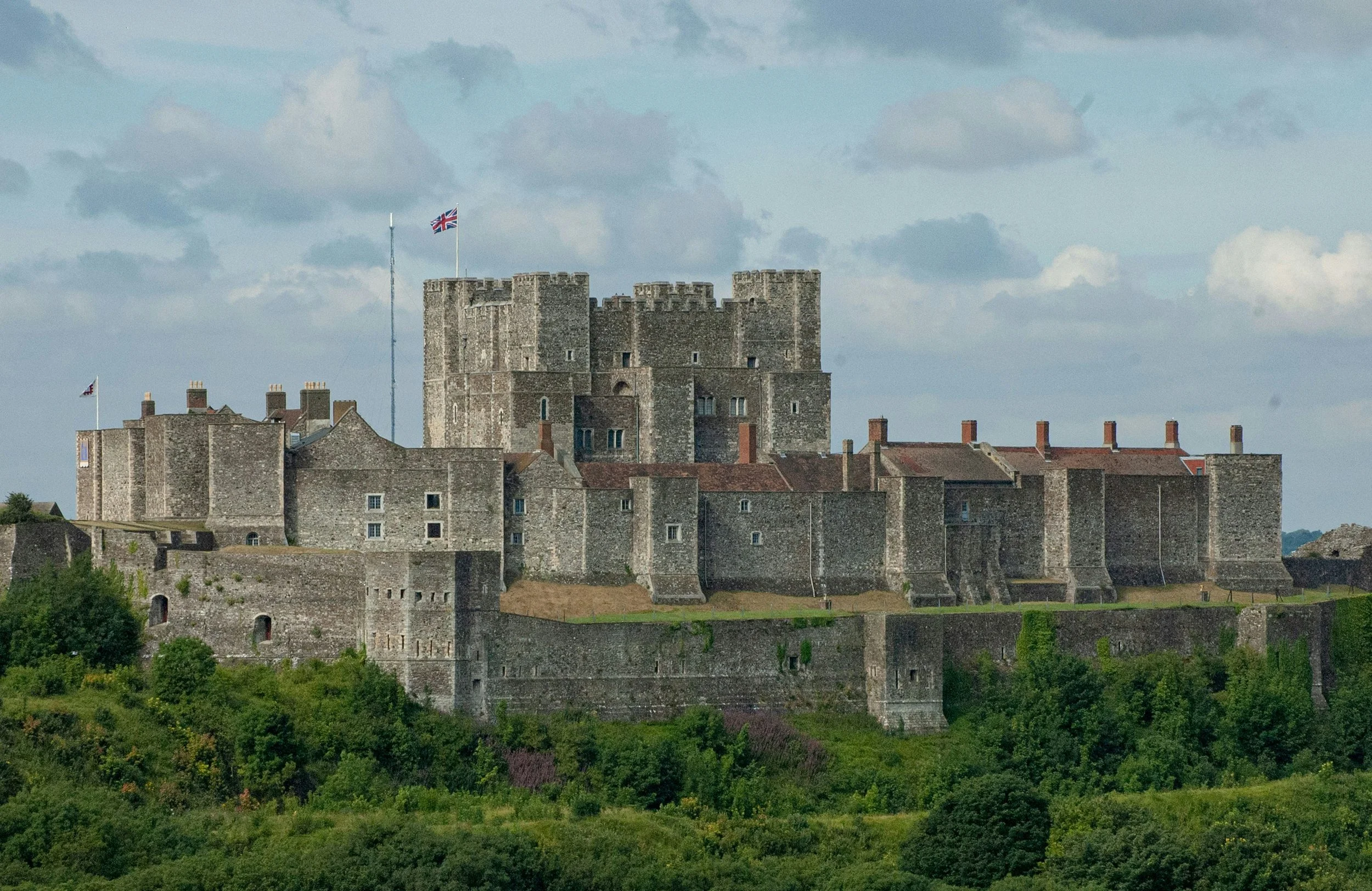 Dover Castle perched on a green hilltop in Kent, with stone walls, central keep, and a Union Jack flag flying under a cloudy sky.