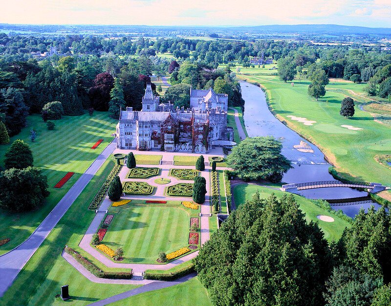 Aerial view of Adare Manor in County Limerick, Ireland, featuring an elegant neo-Gothic mansion surrounded by formal gardens, lush green lawns, a river, and a beautifully landscaped golf course.