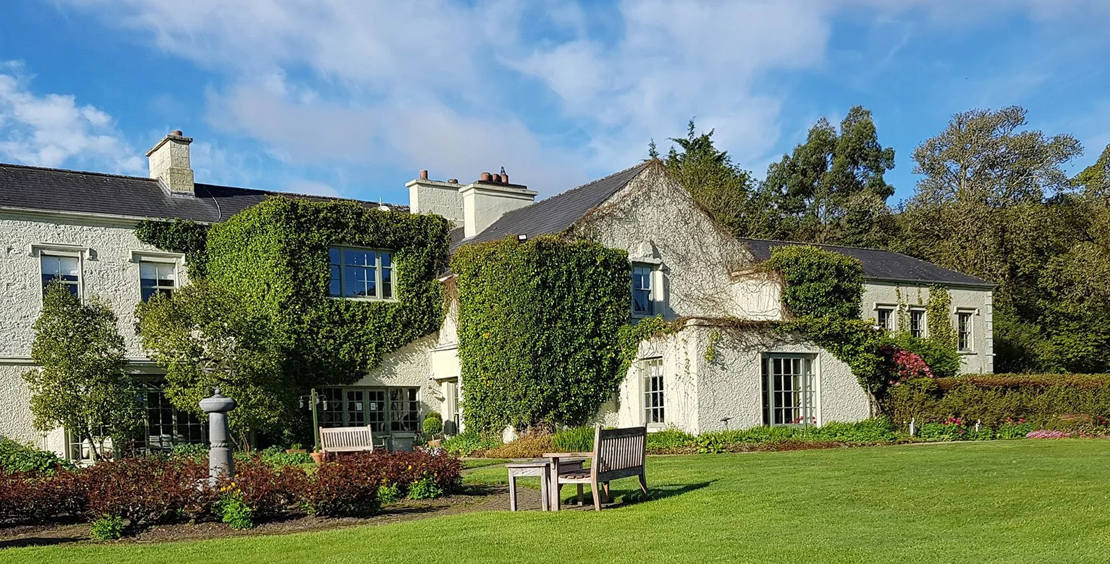 Gregan's Castle Hotel, a white-rendered Irish country house hotel with ivy-covered facade, slate roof and multiple chimneys, set against a manicured garden with wooden benches, colourful borders, and mature trees under a bright blue sky.