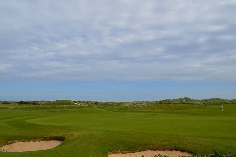Expansive view of the green at Royal Portrush Golf Club in County Antrim, featuring a neatly trimmed fairway, sand bunkers in the foreground, rolling dunes in the distance, and a partly cloudy sky overhead.