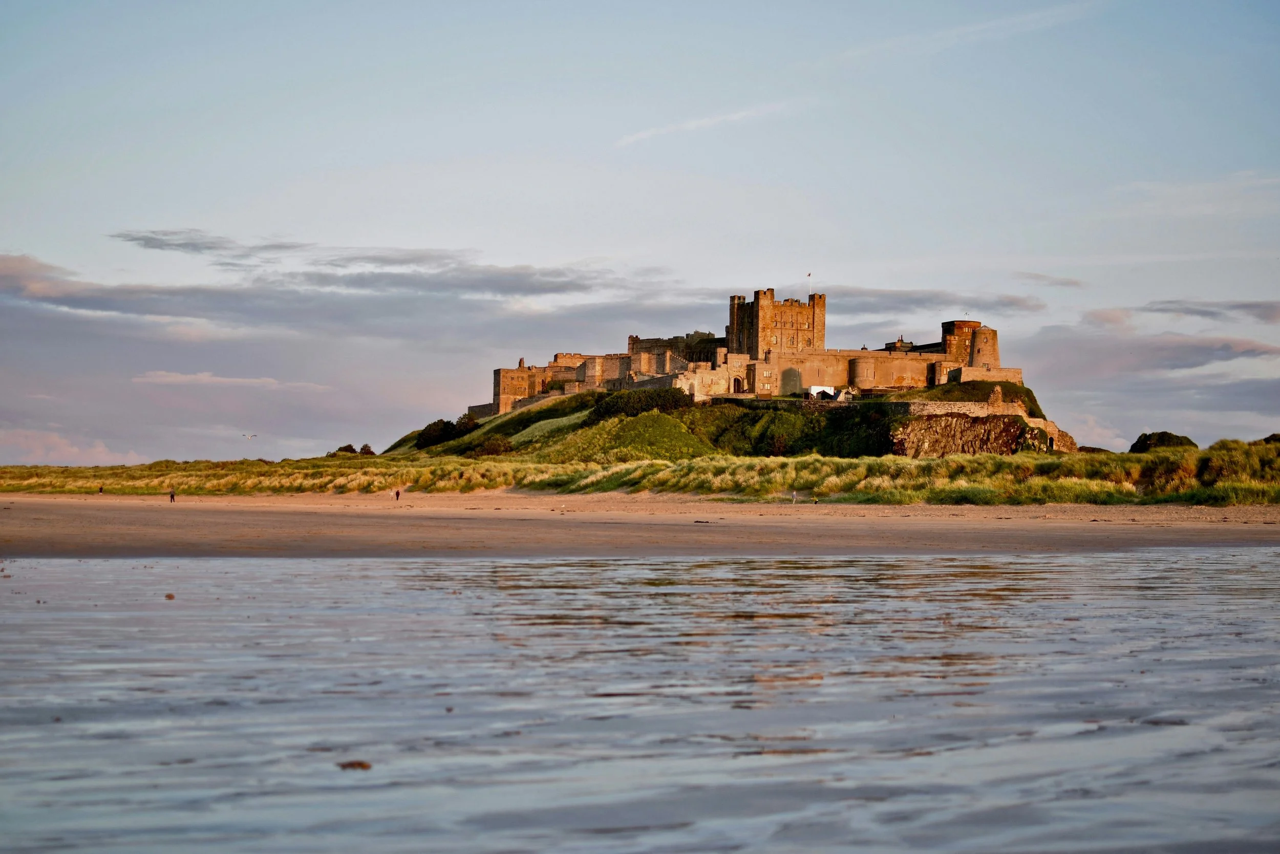 Bamburgh Castle in Northumberland glowing in the evening light above grassy dunes, viewed from the sandy beach with calm water in the foreground.