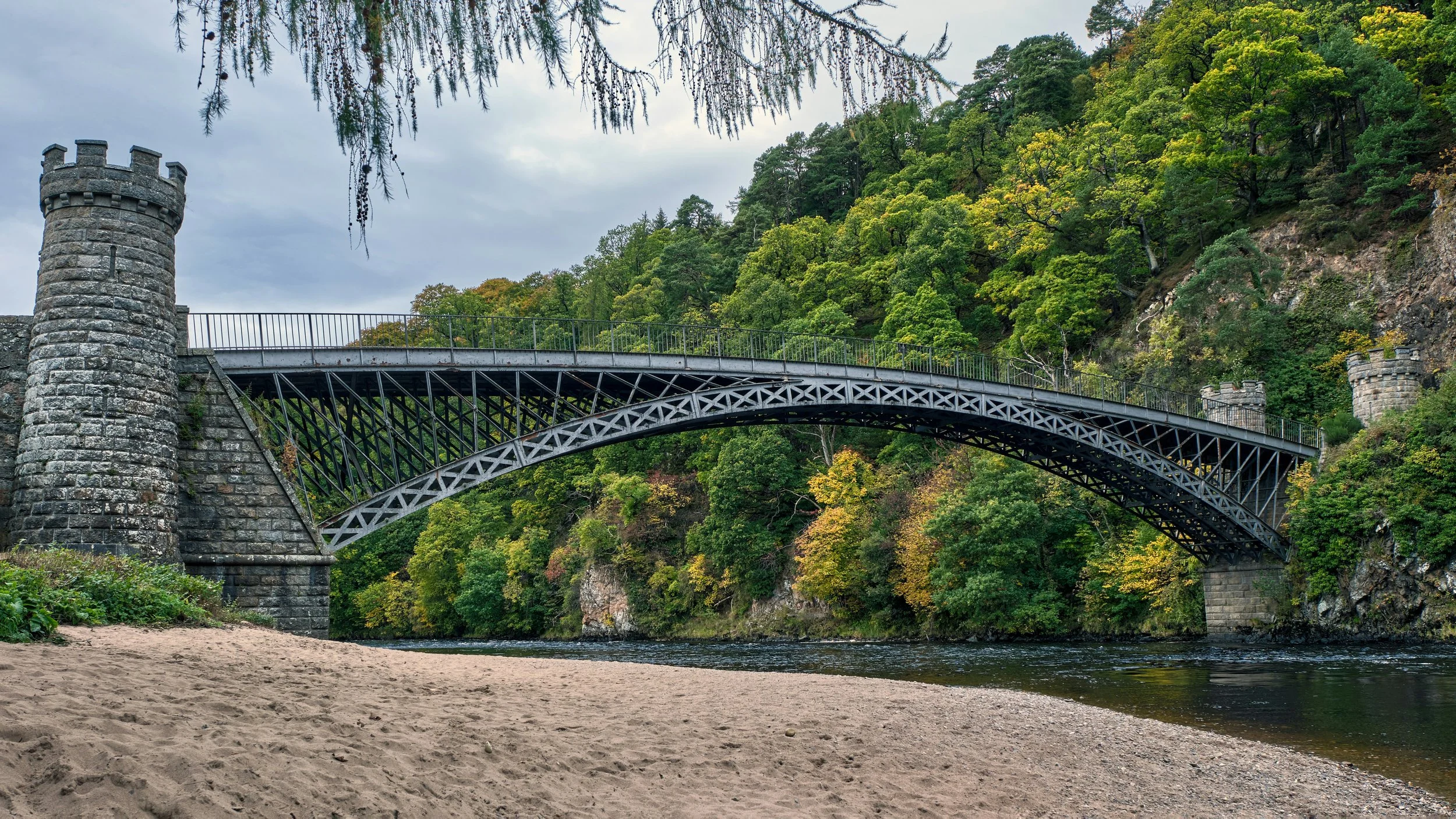 Iron arch bridge with stone towers spanning a river in a forested valley, viewed from a sandy riverbank under an overcast sky.