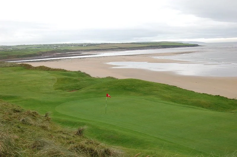 Scenic view of a coastal golf course overlooking a sandy beach with rolling green hills and a red flag marking the hole at Ballybunion Golf Club in Ireland.