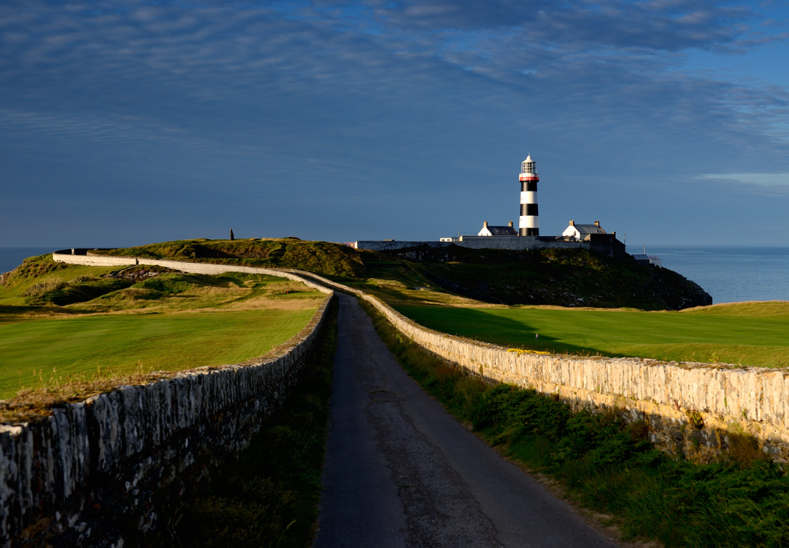 A narrow road lined with stone walls leads toward a black-and-white striped lighthouse perched on a cliff overlooking the sea at Old Head Golf Links in Kinsale, Ireland, under a partly cloudy sky.