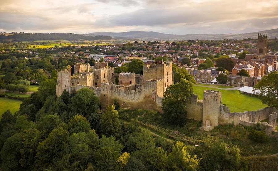 Aerial view of Ludlow Castle in Shropshire, showing its stone ruins, central tower with a flag, and surrounding green countryside under a cloudy sky.