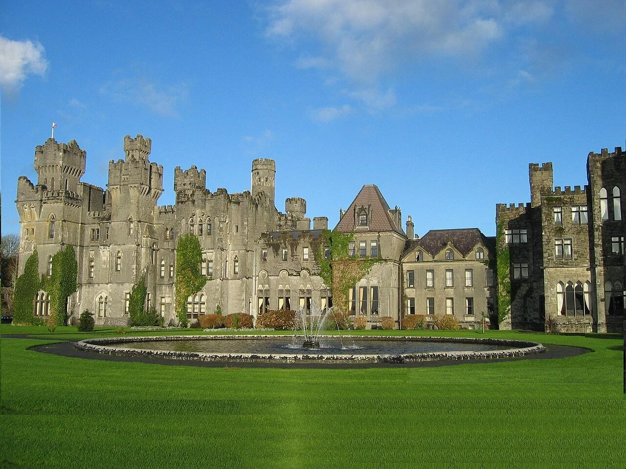 Ashford Castle, a medieval-style stone castle hotel in County Mayo, Ireland, featuring battlemented towers, Gothic arched windows, ivy-covered walls, and a circular fountain on a manicured green lawn, under a blue sky with scattered clouds.