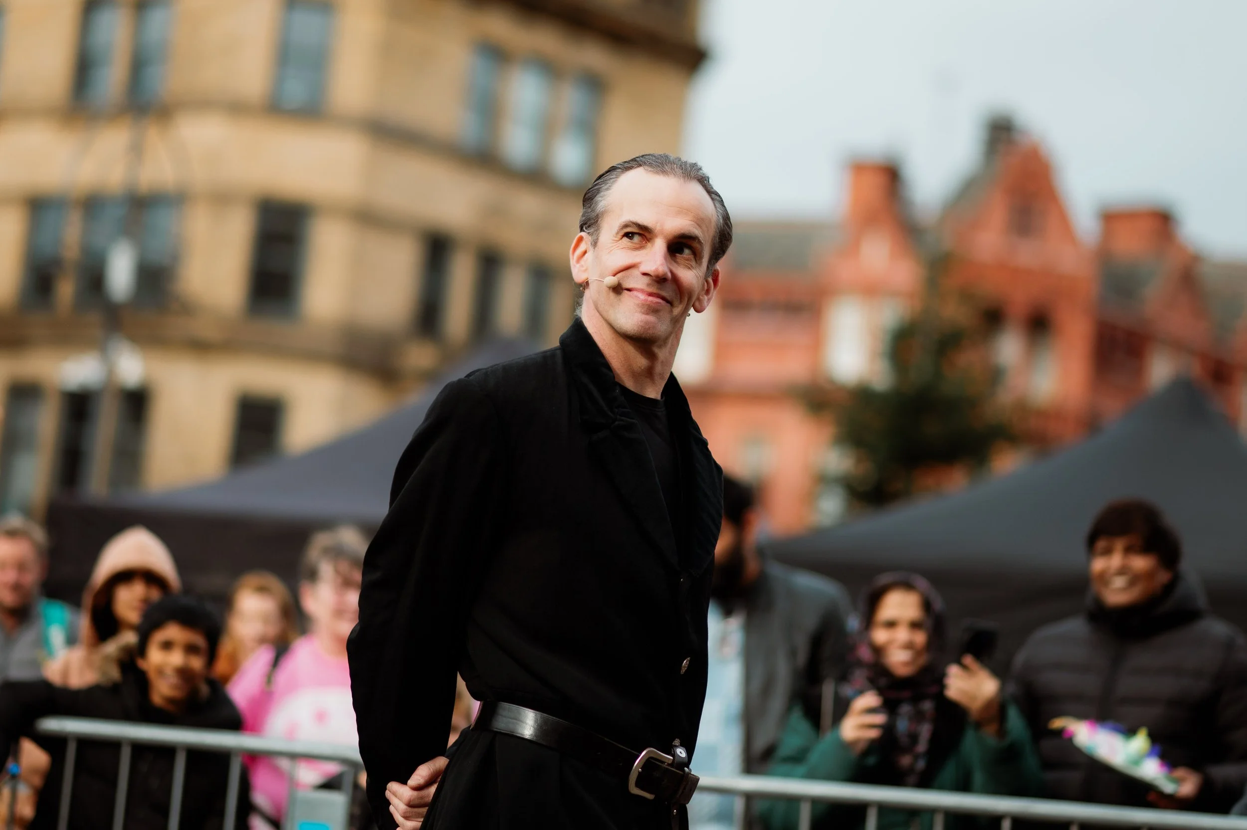 A man with dark hair, dressed in black, stands with a slight smile in front of a group of people at an outdoor event. The background features buildings with distinct architecture and tents.