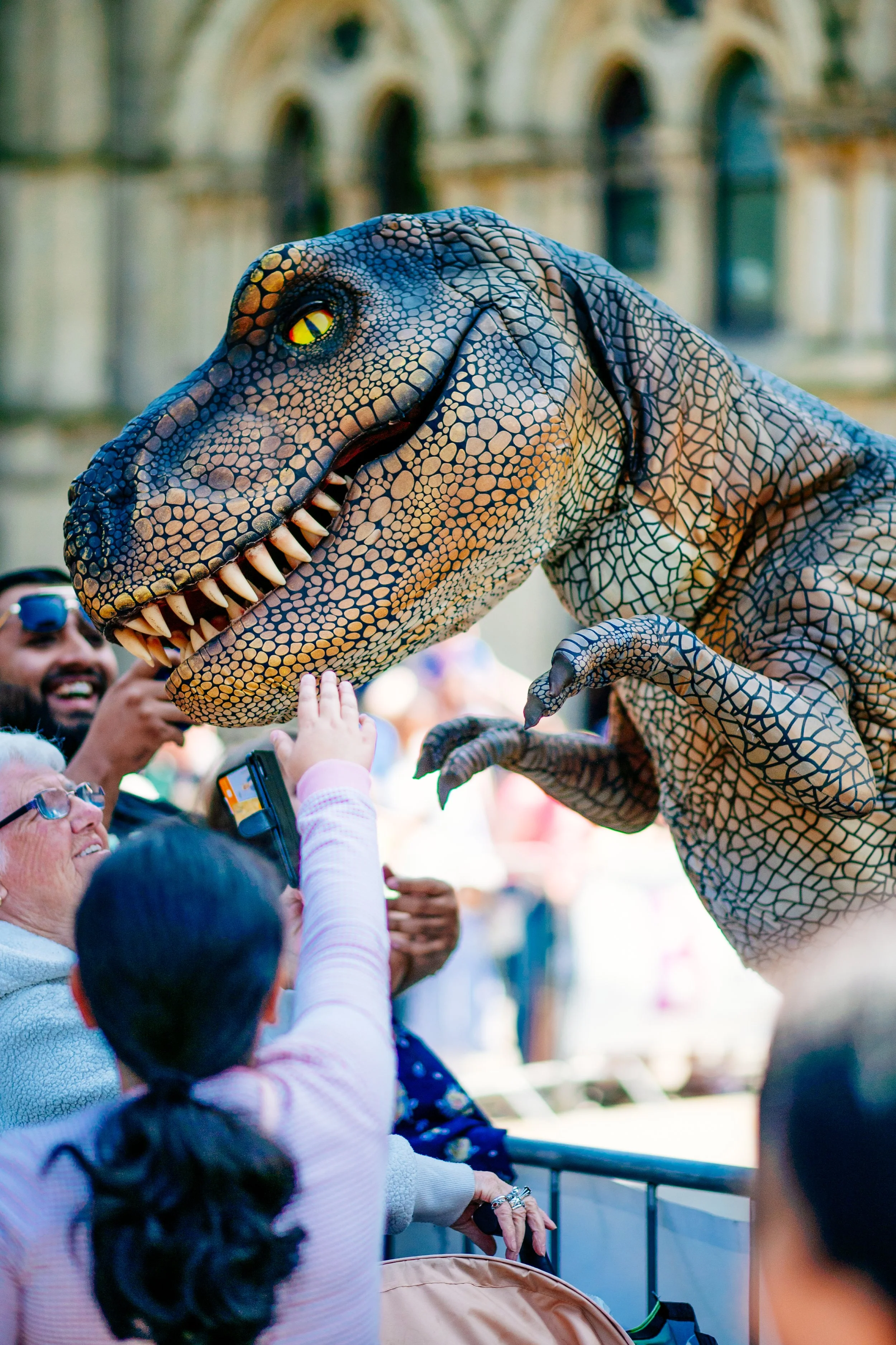 A person in a dinosaur costume high-fives children during a public event with a historic building in the background.