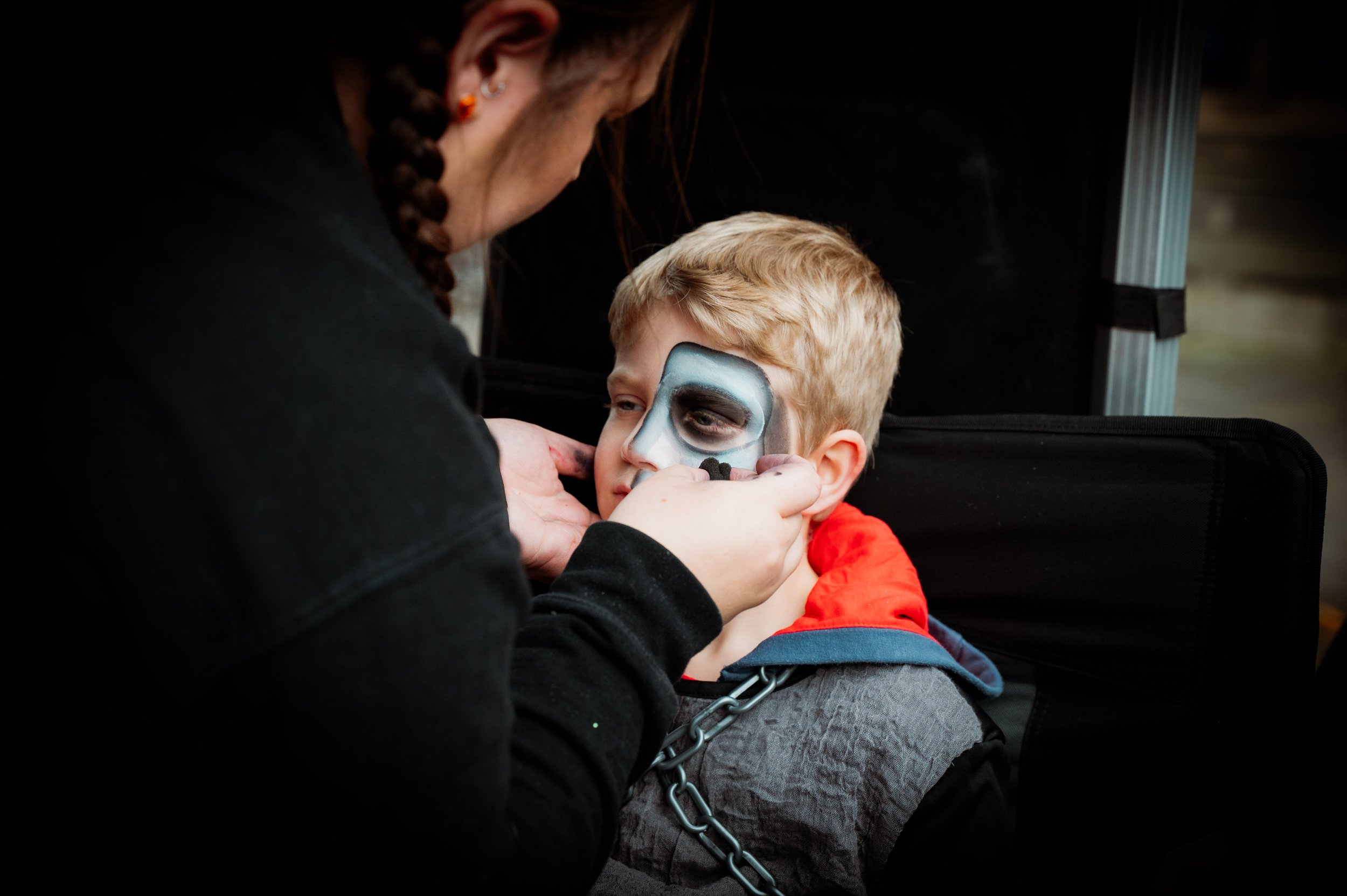 A woman is applying face paint to a young boy's face, who has blonde hair and is wearing a gray vest with a red hoodie underneath. The boy's face has a partially painted design, and he is sitting in a black chair while the woman, dressed in black wit
