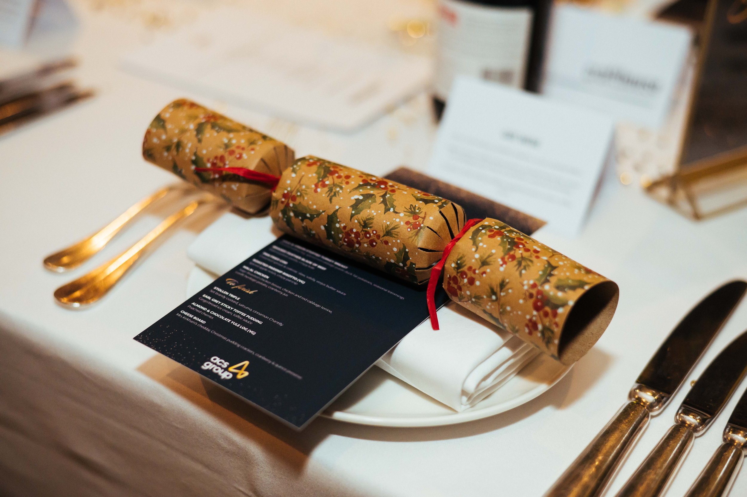 Festive holiday table setting with a scroll of wrapping paper, a black menu card, white folded napkin on a white plate, gold cutlery, and blurred background with additional table decor.