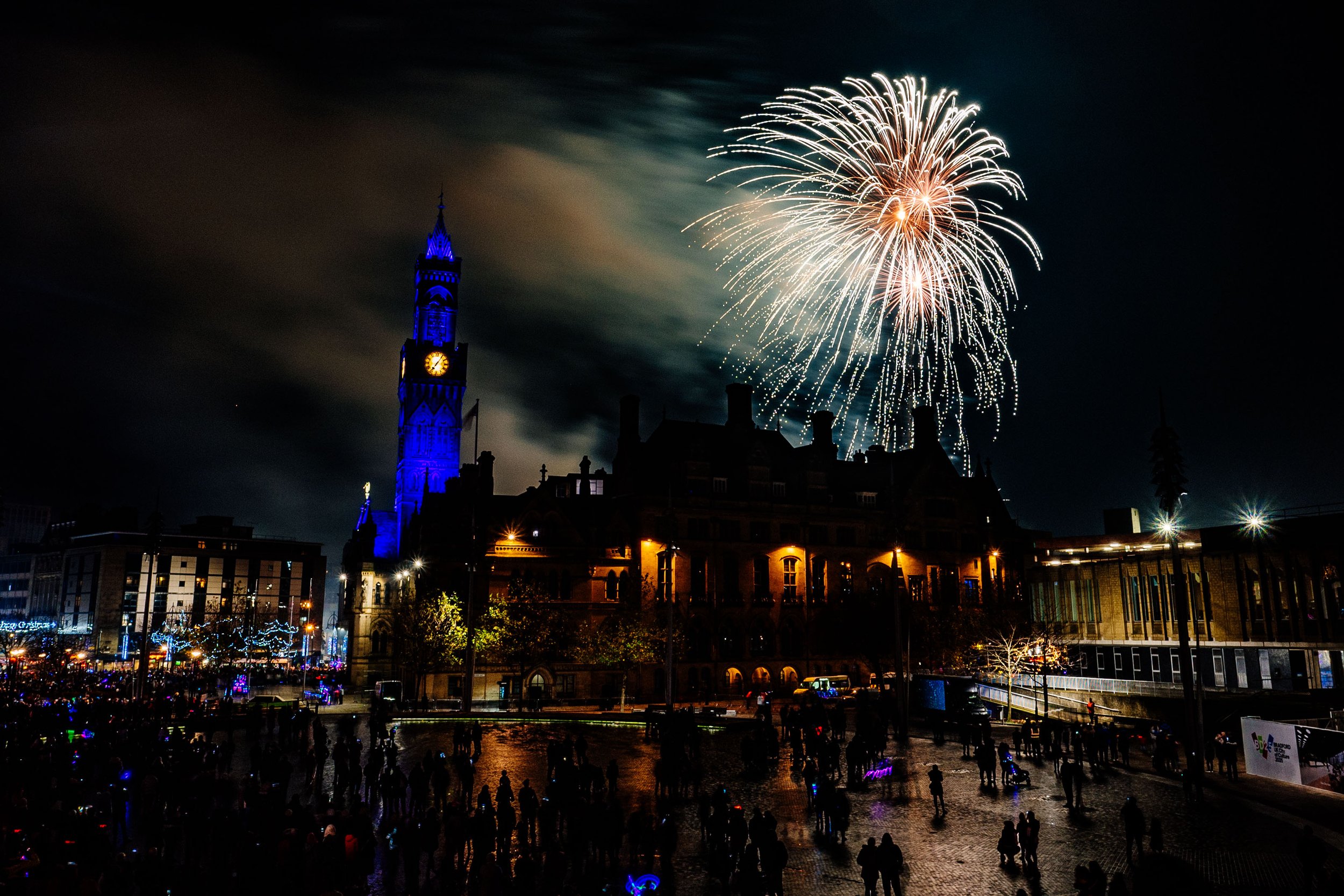 Nighttime fireworks display over a city with a clock tower illuminated in blue, crowds of people in the foreground celebrating.