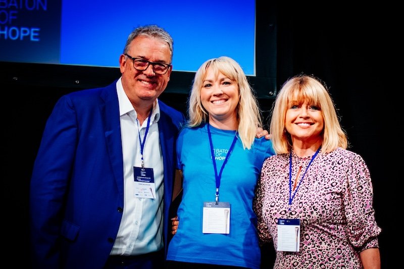 Three smiling people posing together at an event, wearing conference badges, with a blue screen or banner in the background.