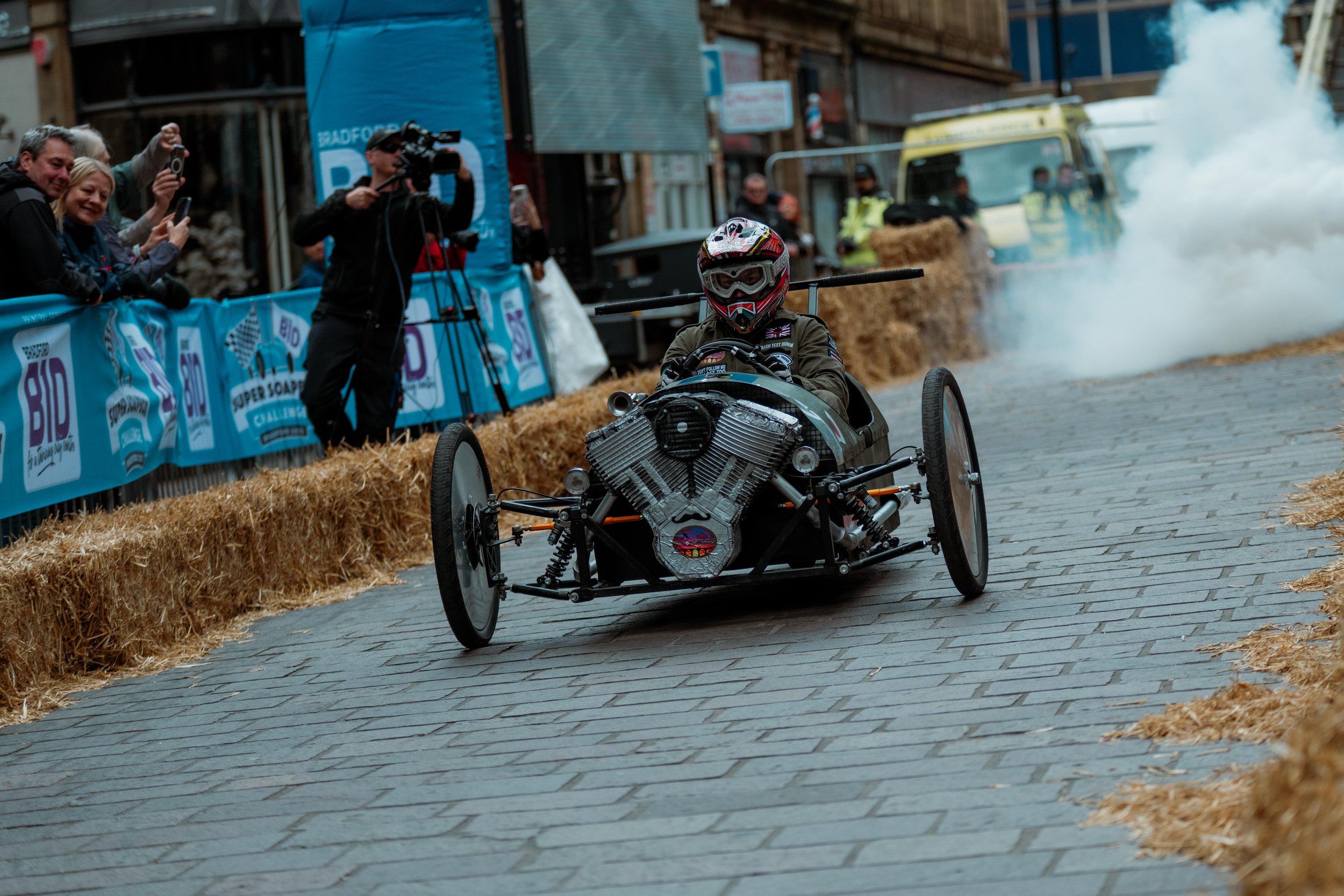 A person in a racing suit and helmet driving a vintage-style racing vehicle on a cobblestone street during a race, with spectators and photographers watching behind hay barriers and smoke in the background.