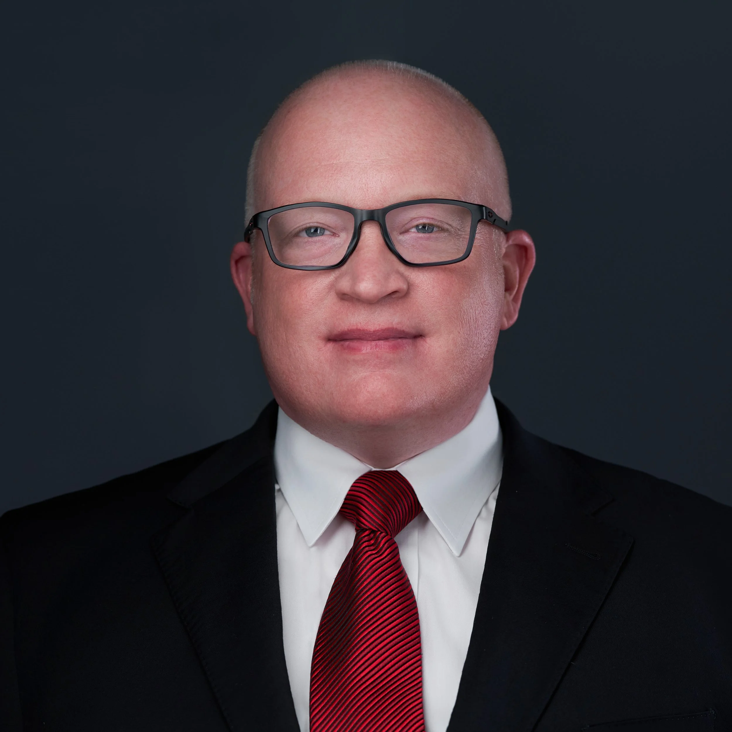 Headshot of a bald man wearing glasses, a black suit, white shirt, and red tie against a dark background.