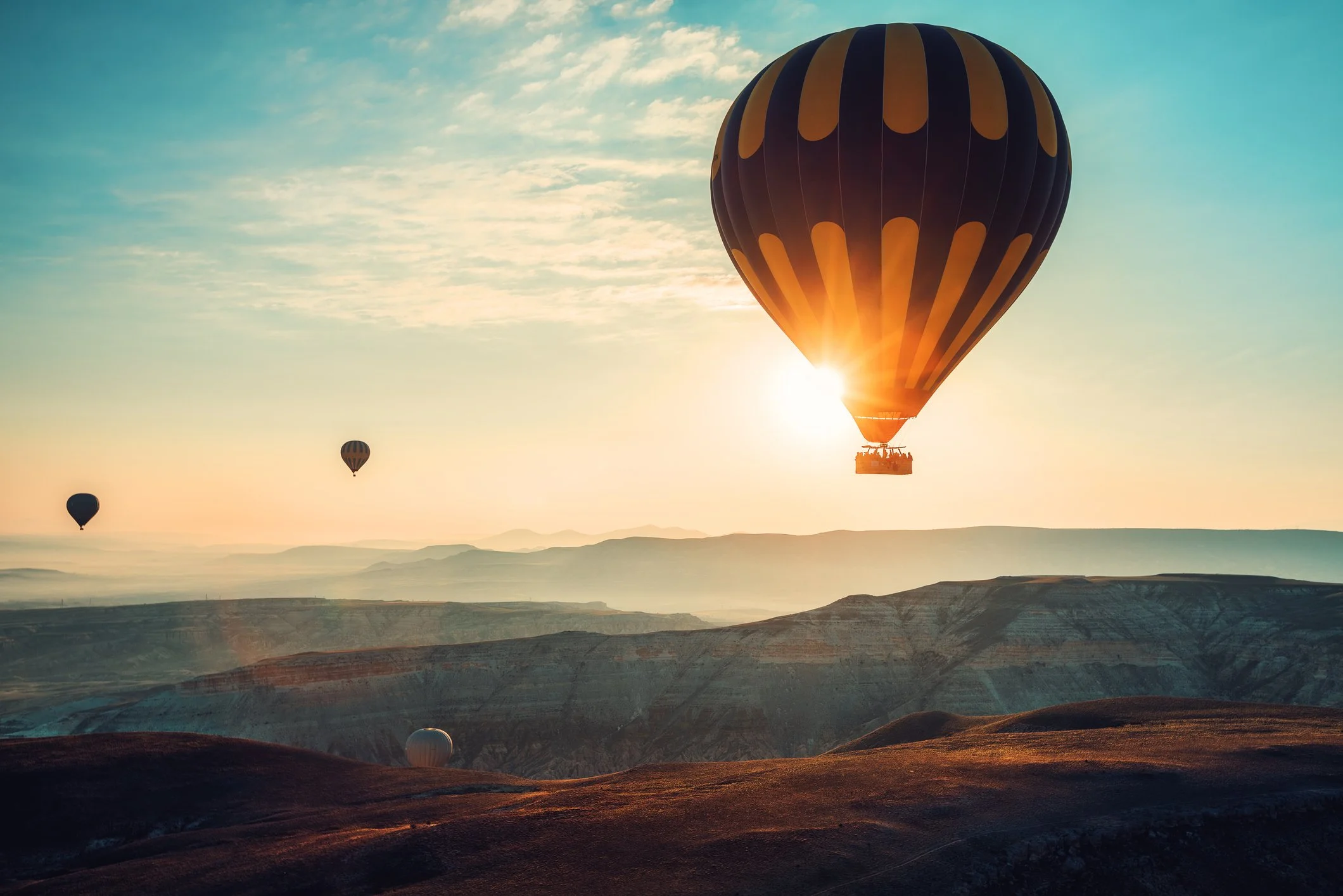Vliegende ballonnen boven een berglandschap bij zonsopgang, met één bal met oranje en zwarte strepen die het zonlicht blokkeert.