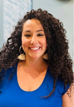 A woman with curly hair smiling and wearing a blue top and large tassel earrings.