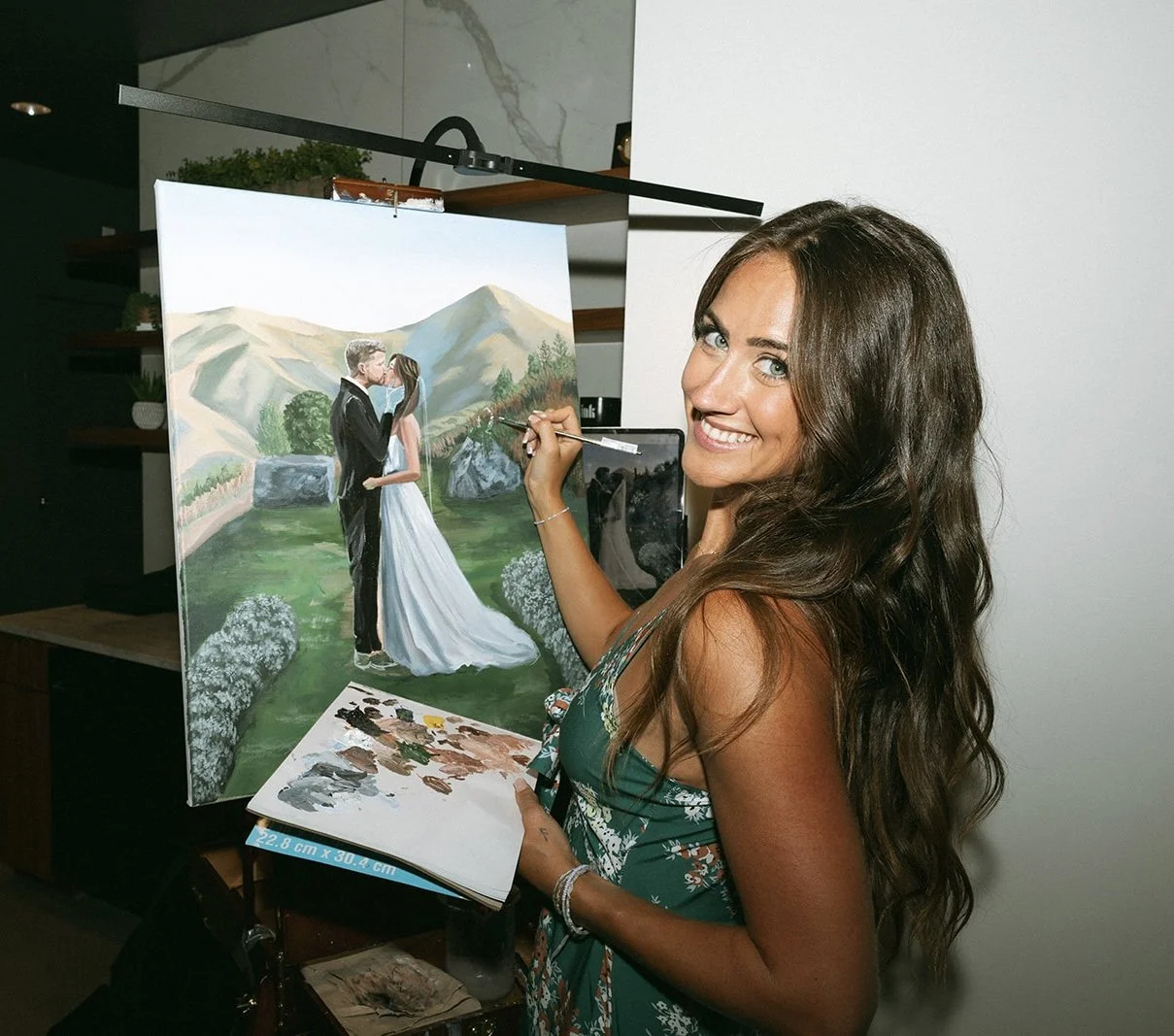 A woman with long, wavy brown hair and a green patterned dress is painting a wedding portrait on a canvas. She is smiling and holding a paintbrush near her shoulder. The painting depicts a couple in wedding attire, standing outdoors with mountains and greenery in the background.