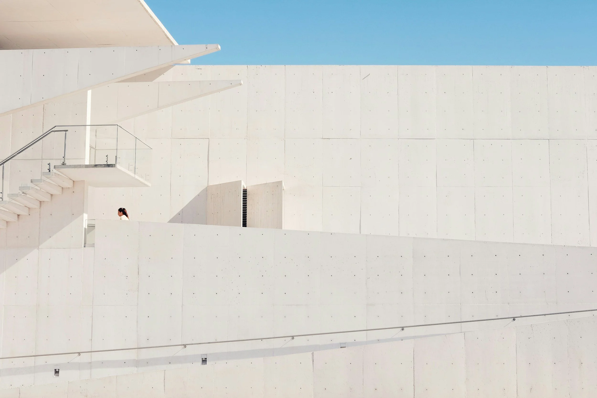 Minimalist white building with stairs and a person standing on a balcony, under a clear blue sky.