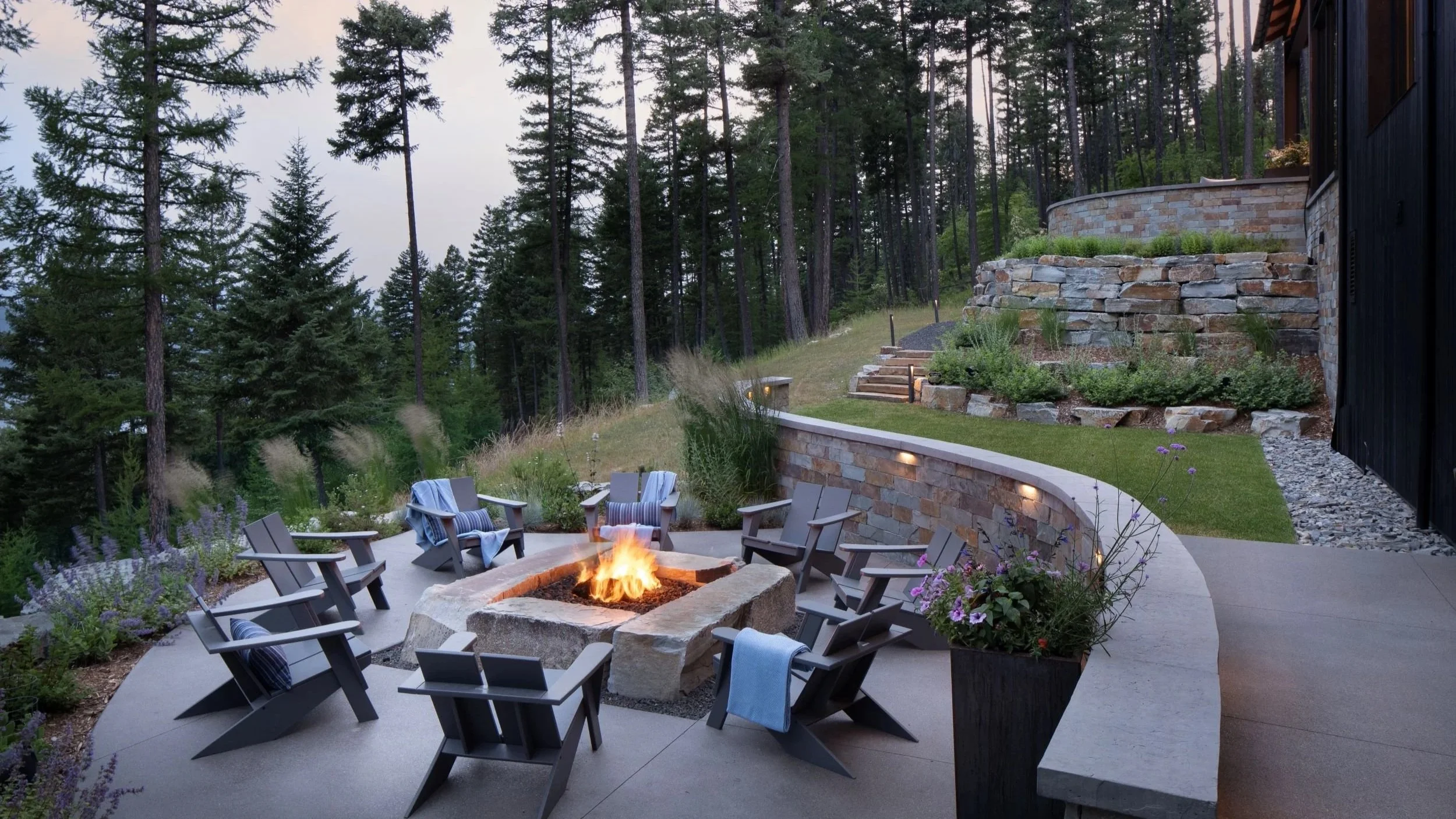 Outdoor patio with a fire pit surrounded by chairs, overlooking a forested landscape with tall trees, a stone retaining wall, and a seating area illuminated by soft lighting.