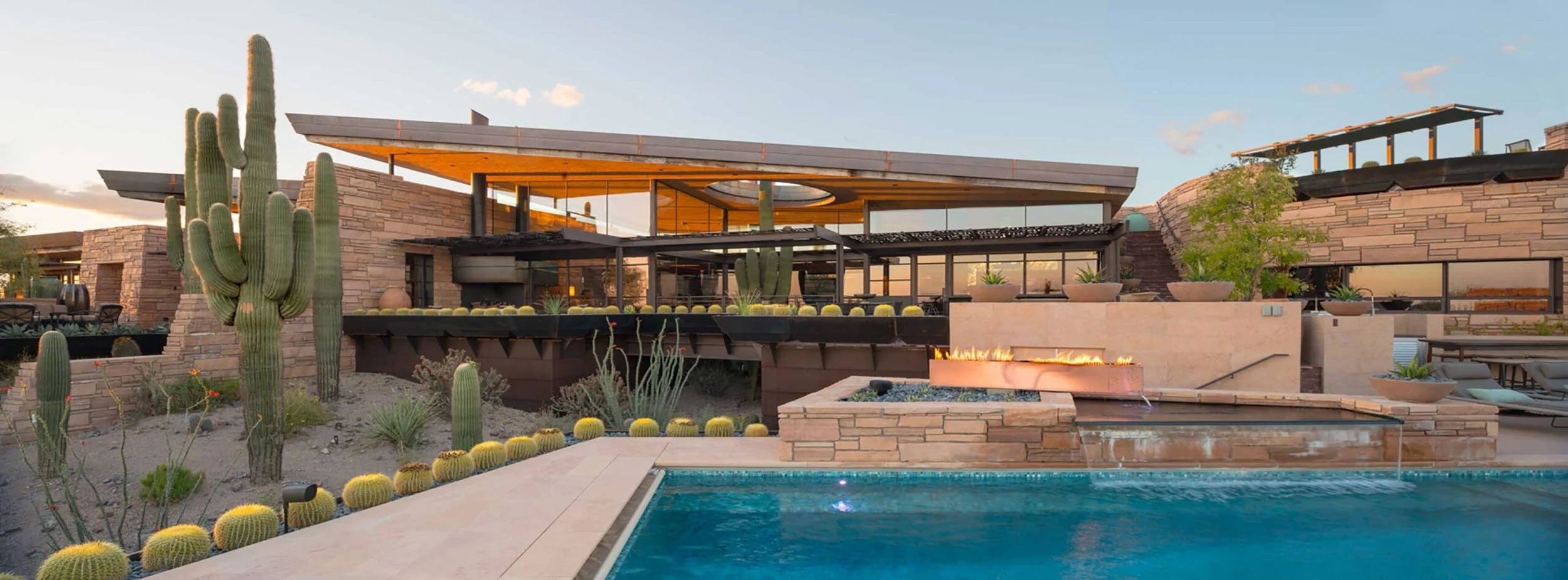 Modern desert-style house with a swimming pool, cacti, and outdoor fire pit at sunset.