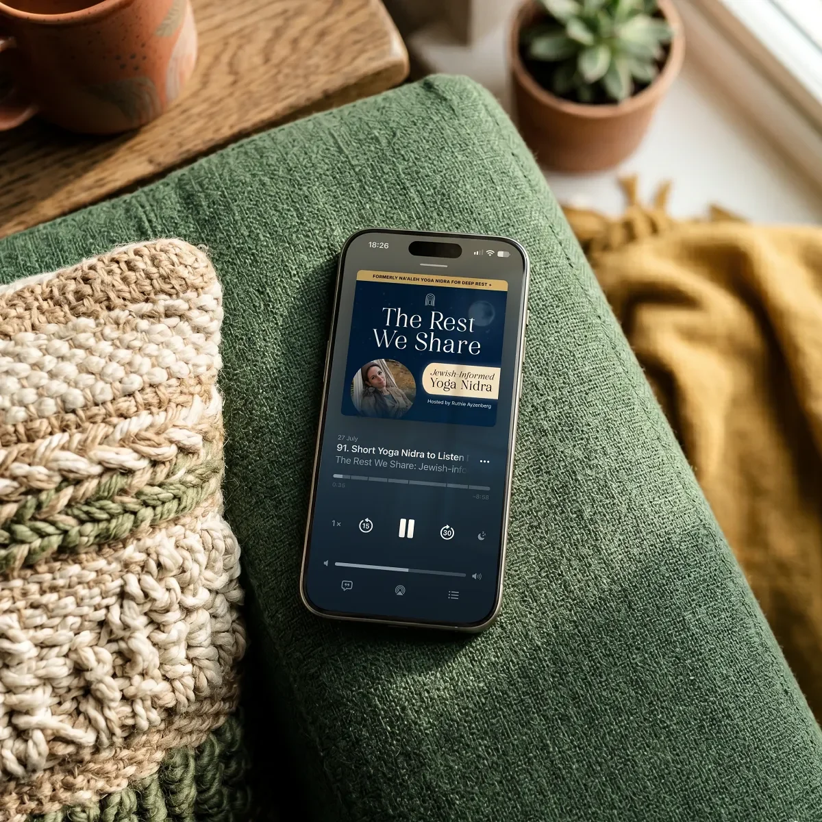 Phone lying on a sofa displaying the podcast The Rest We Share: Jewish-Aligned Yoga Nidra by Ruthie Ayzenberg.