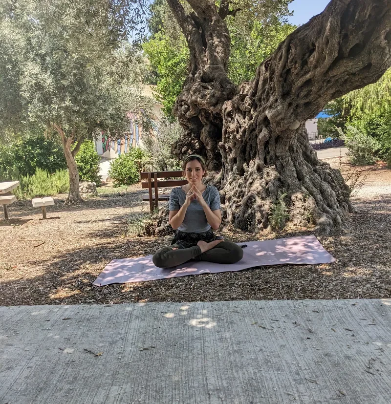 Ruthie Ayzenberg seated outdoors on a yoga mat beneath an olive tree.