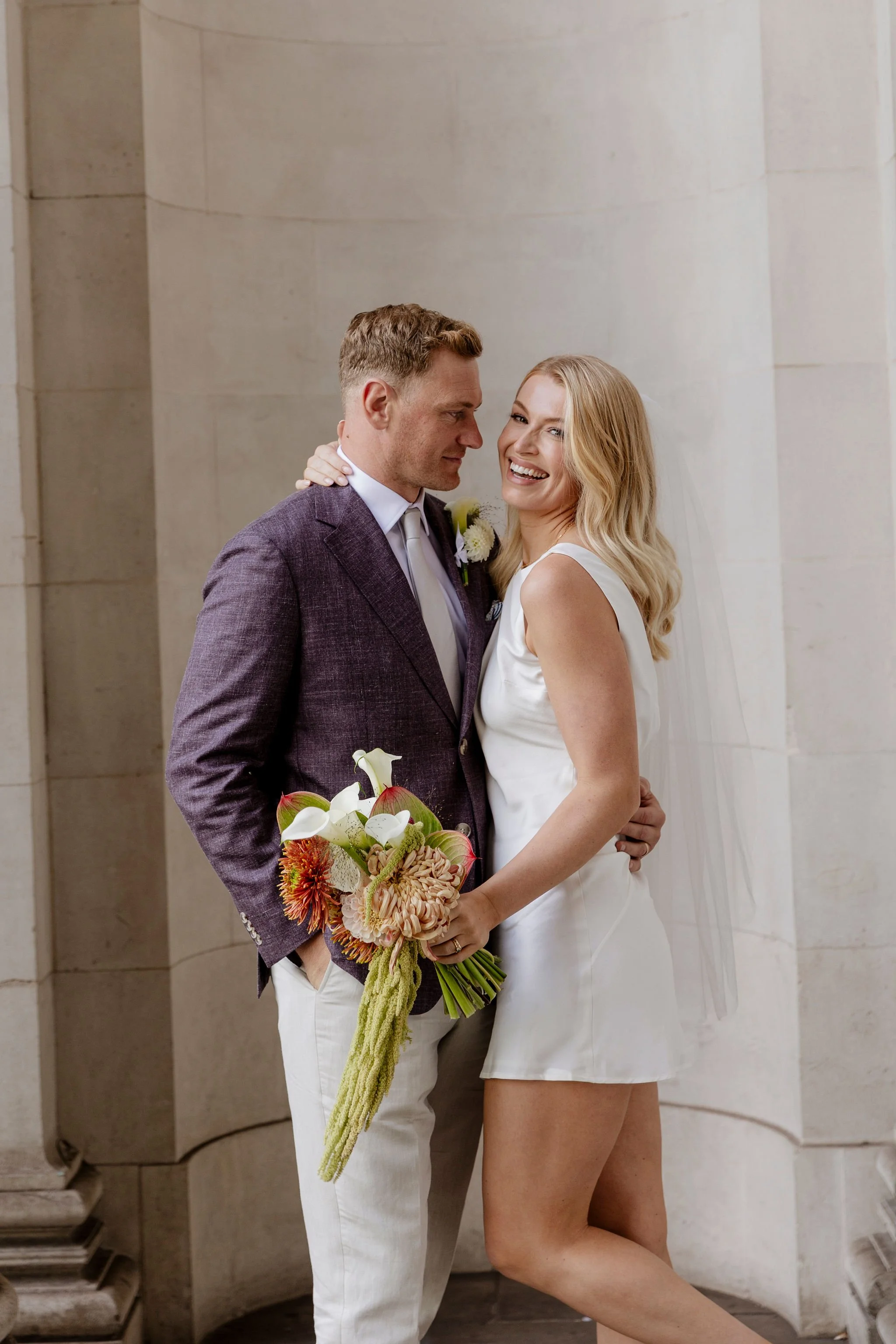 Elegant city-hall wedding couple. The bride holds a modern, sculptural bouquet of calla lilies and trailing amaranth, designed in a minimalist contemporary style. Minimal small bouquet.