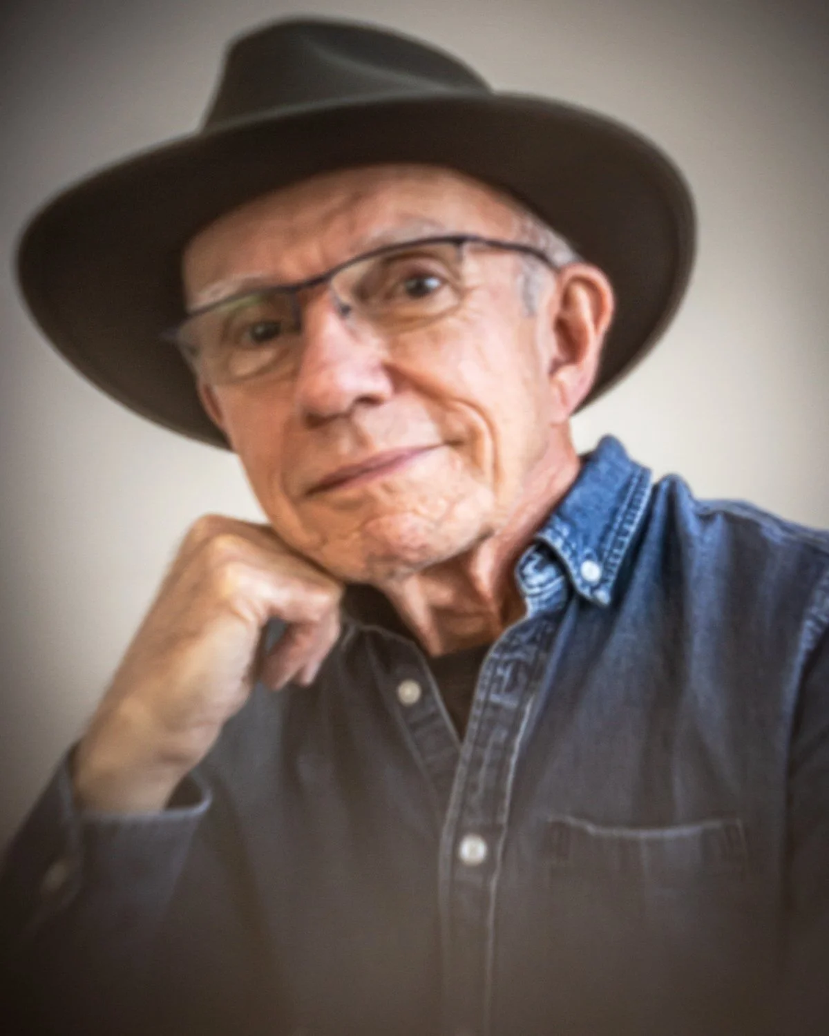 A man with glasses, a black hat, and a black shirt standing against a dark wall.
