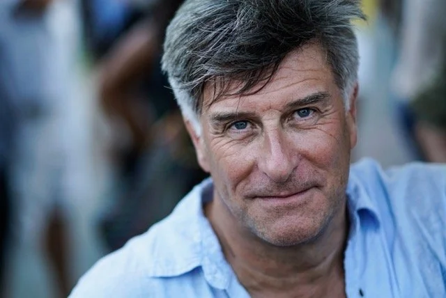 Close-up of a middle-aged man with gray hair, wearing a light blue collared shirt, smiling gently with a blurred background.