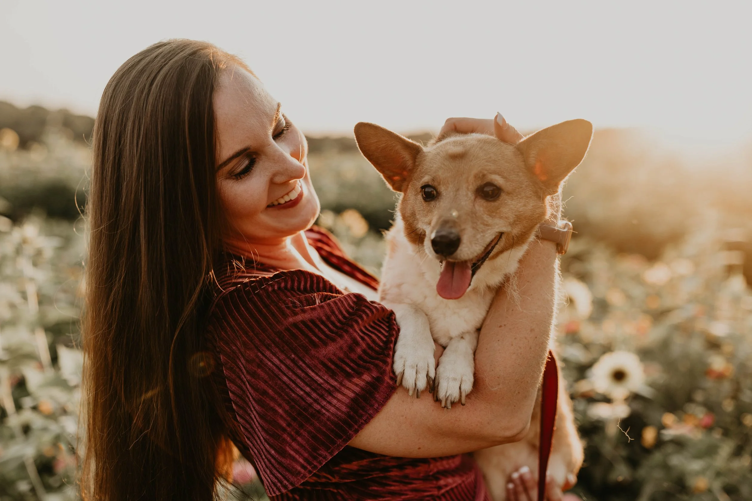 A woman with long brown hair smiling and holding a happy small tan and white dog outdoors during sunset.