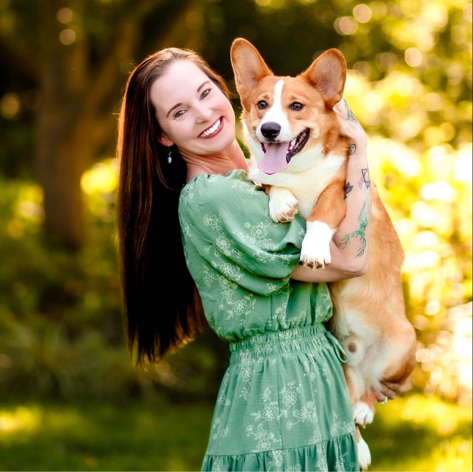 A woman with long brown hair and a green dress holding a happy Corgi dog outdoors during daytime.