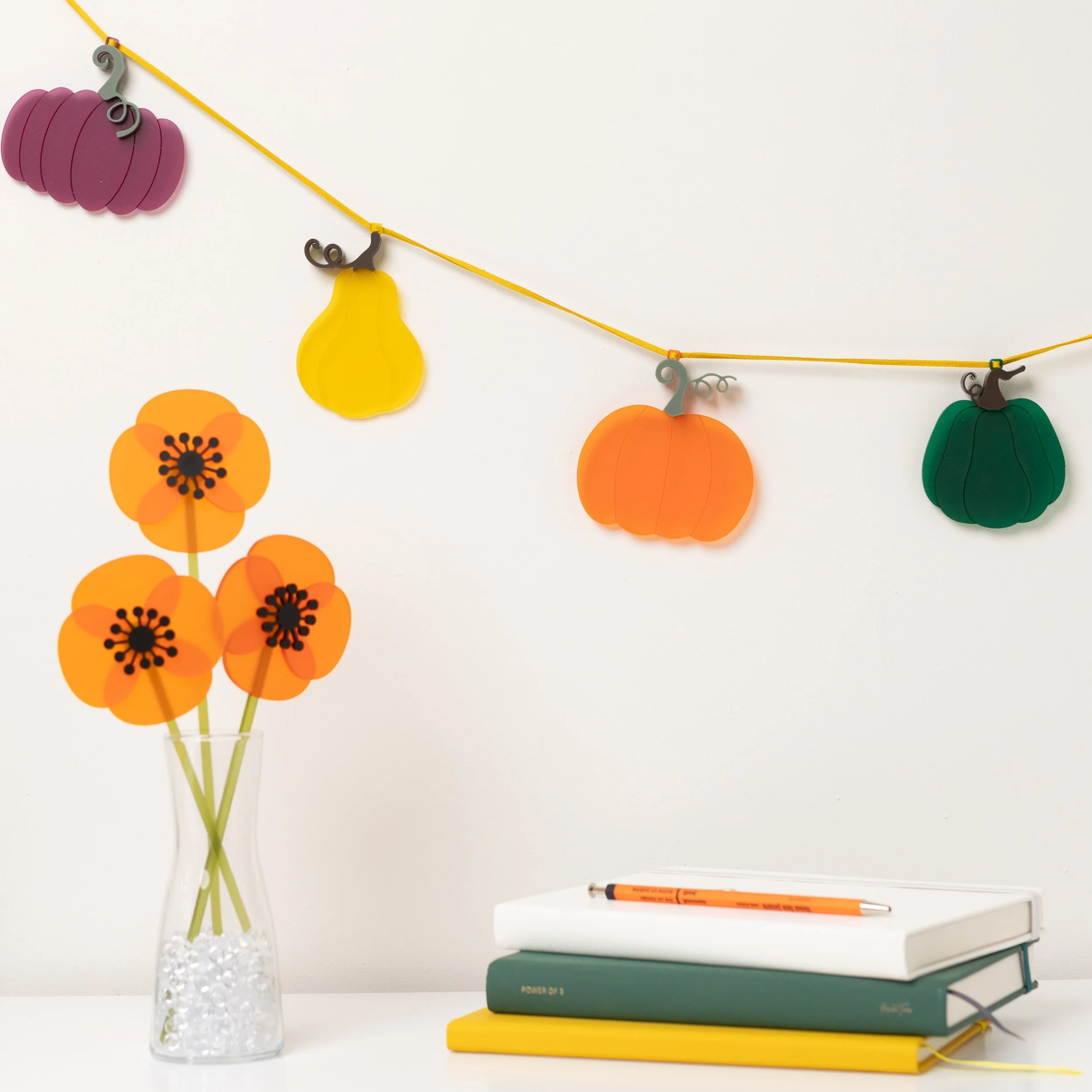 A squash garland showing 4 of 9 squashes on a white wall. Theres' an orange, plum, saffron & emerald pumpkin that are all different shapes. On the desk is a vase of acrylic orange poppies and a pile of books