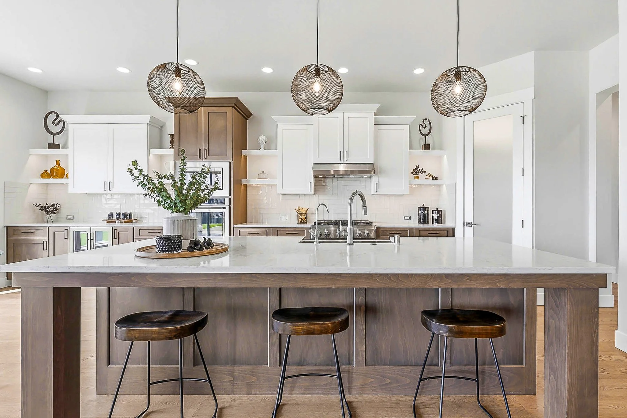 Modern kitchen with white cabinets, a large island with a wooden base, and three pendant lights hanging above. There are three dark wood barstools at the island and some decorative items on the shelves.