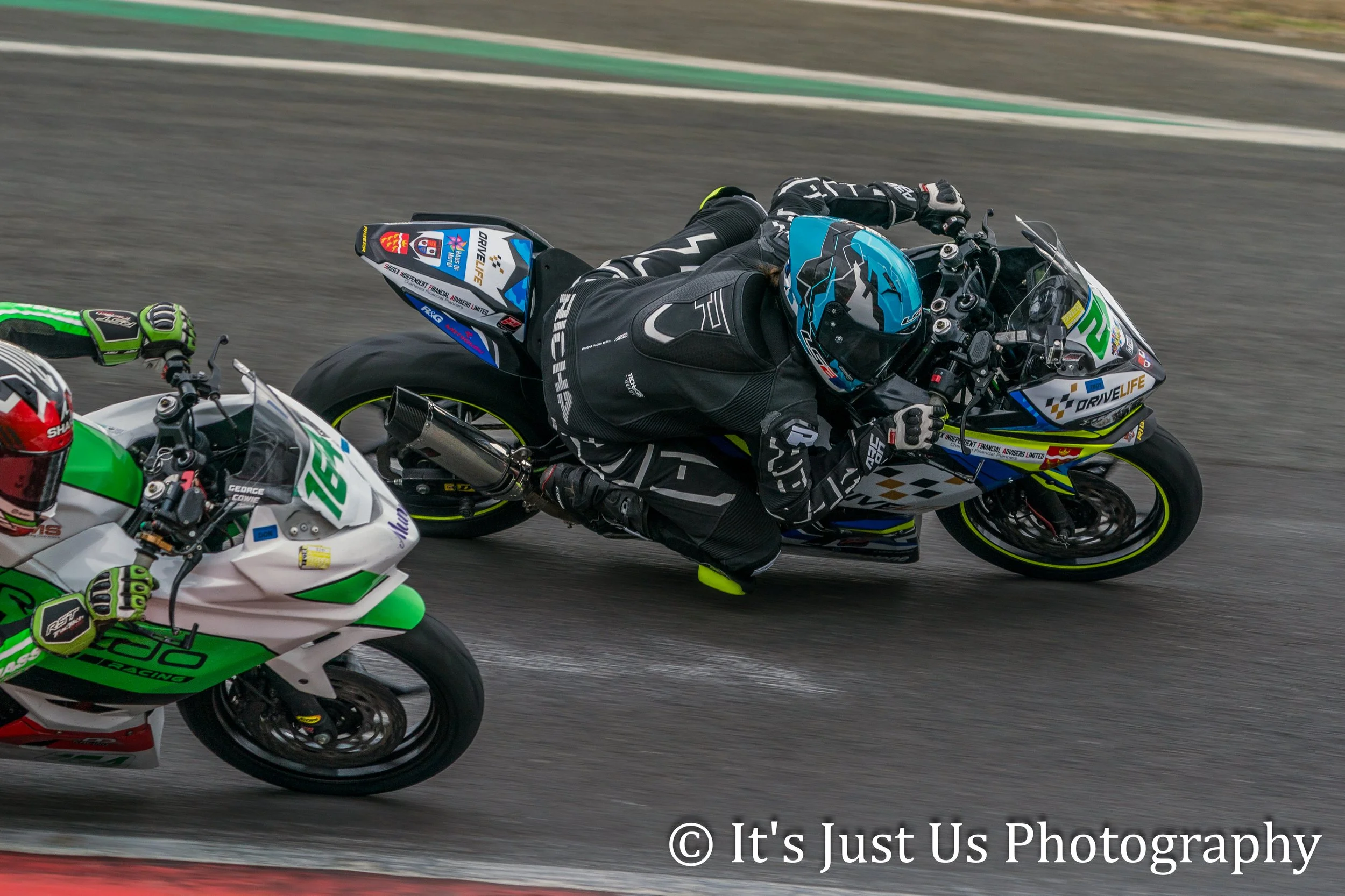 Two motorcycle racers leaning into a turn on a race track, one wearing a black suit with a blue helmet, and the other in a white and green suit with a red helmet.