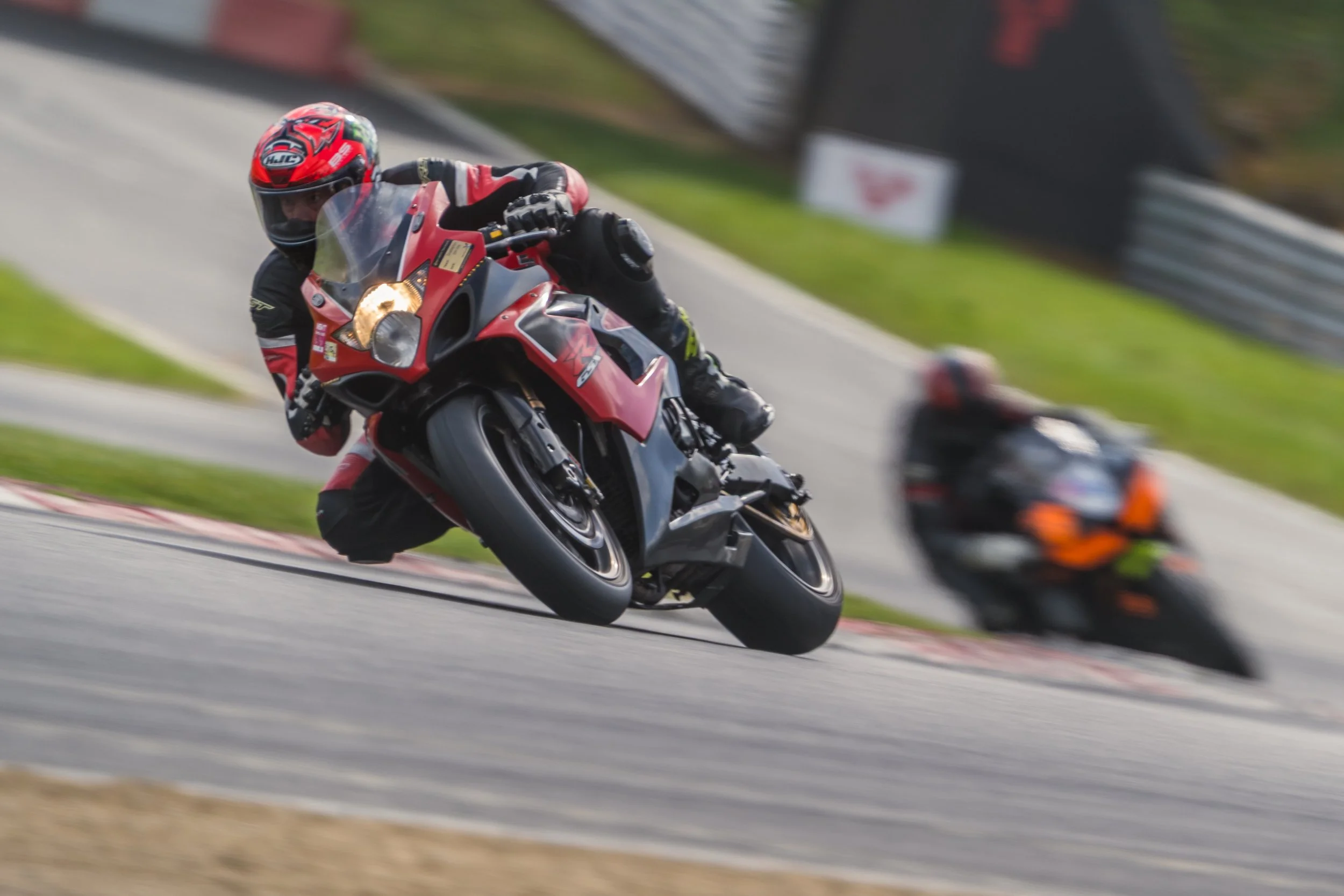 Two motorcyclists leaning into a turn on a racing track, with the rider in the foreground wearing a red and black helmet and suit, and the rider in the background wearing an orange and black outfit.
