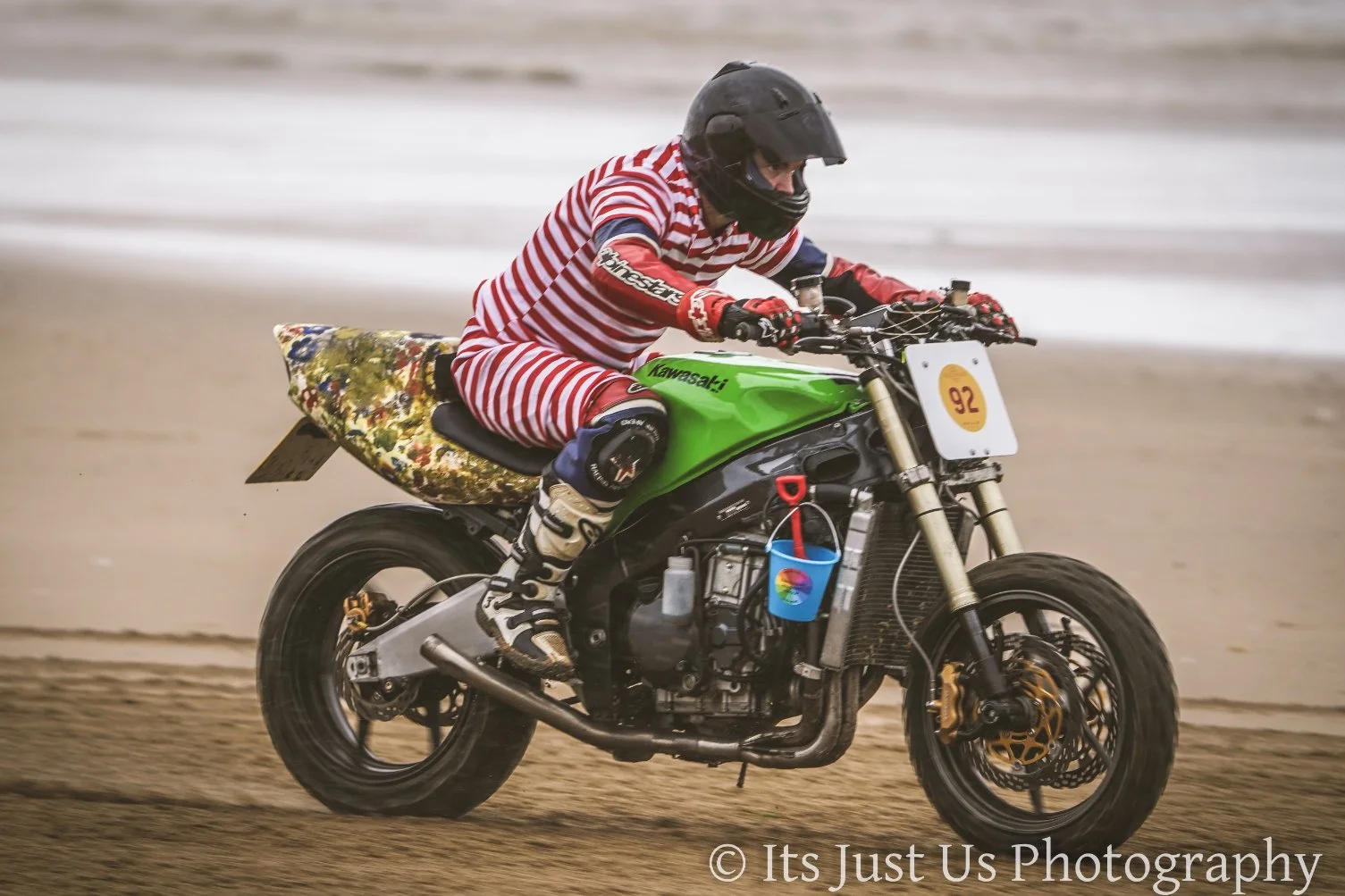 Person riding a green motorcycle on a dirt track, wearing a black helmet and red and white striped outfit.
