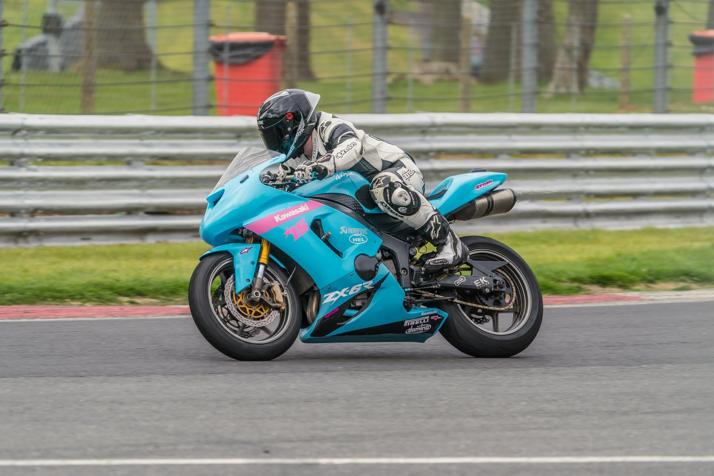A motorcycle racer in a white and black suit riding a blue Kawasaki ZX-6R motorcycle on a race track, with a safety guardrail and trees in the background.