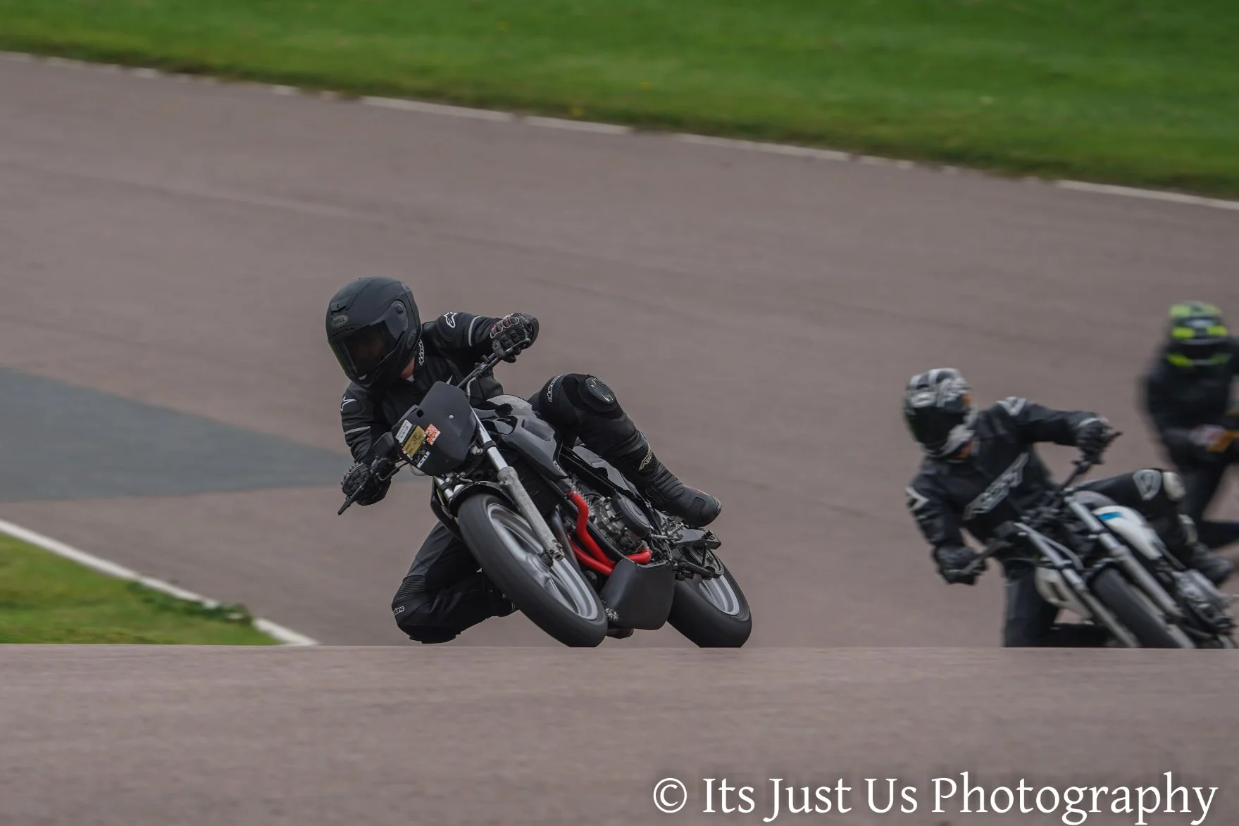 Multiple motorcyclists racing on a track, leaning into a turn. They wear black helmets and gear, with one rider in the foreground on a black and gray motorcycle.