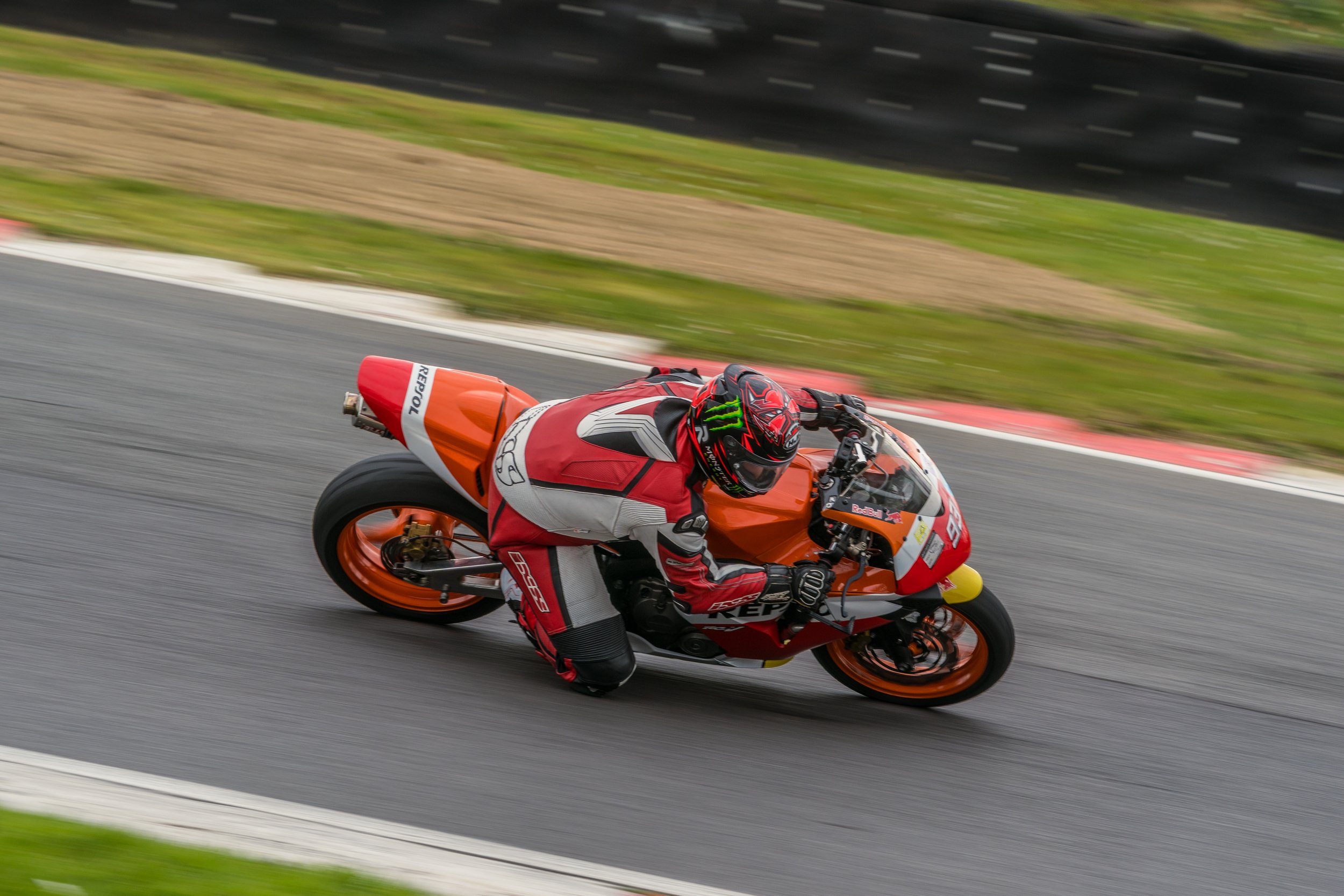 A motorcycle racer in red and black gear riding an orange racing motorcycle on a track with green grass and a black barrier in the background.